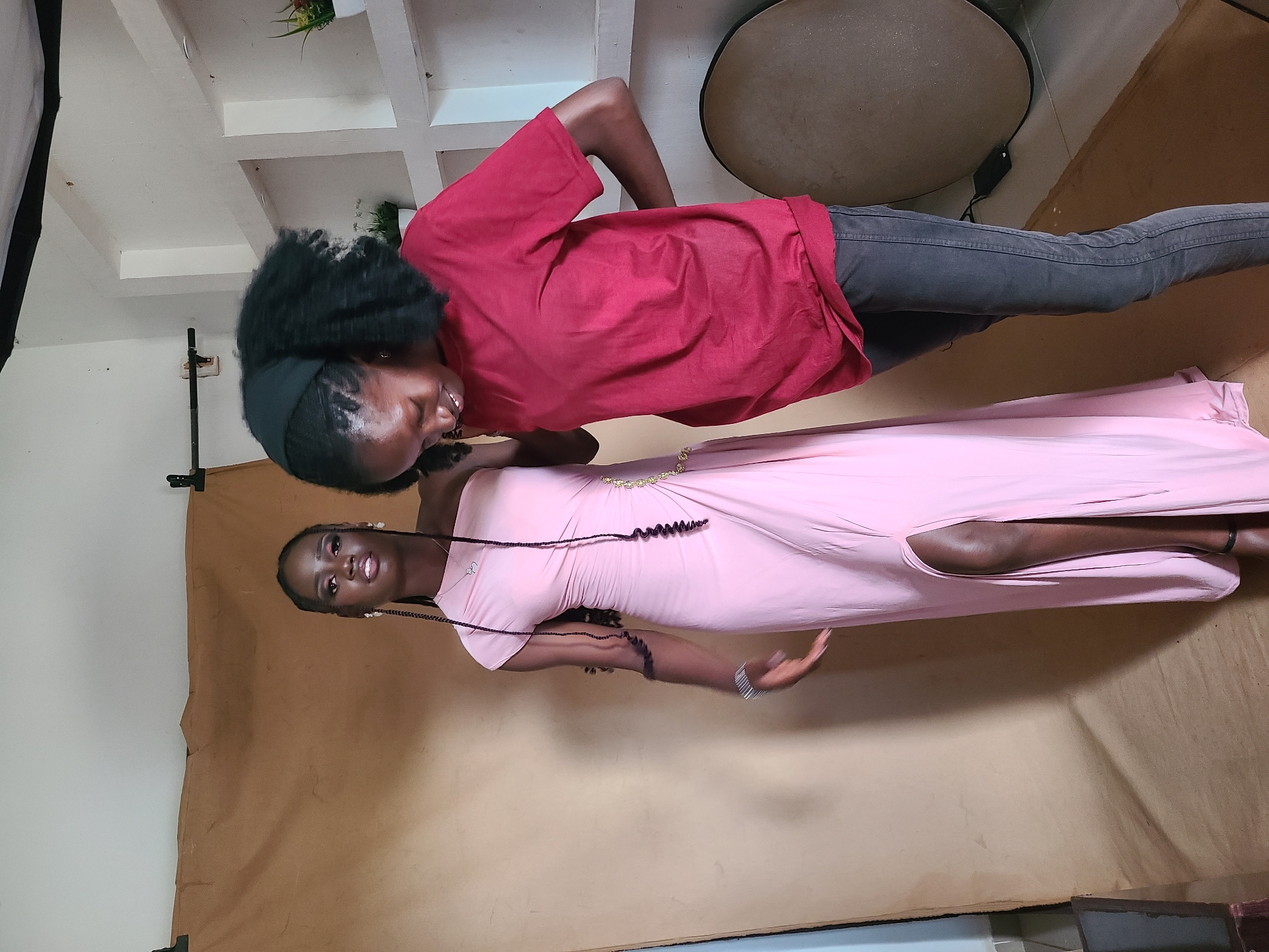 Two young Black women are posed for a photograph indoors. One woman, on the left, is wearing a red t-shirt and dark jeans. She has her hair in a high afro and is wearing a black headband. She is looking down and smiling. The second woman, on the right, is wearing a long, light pink dress with a high slit. Her hair is in braids and she is wearing pearl earrings and a bracelet. She is looking directly at the camera with a neutral expression. The background appears to be a studio setting with a light-colored wall and shelves in the upper left corner. There is a large, round object, possibly a photography light reflector, in the upper right corner. A brown backdrop is behind the women. The lighting suggests it is daytime, but the exact time of day is not discernible. There is no visible text in the image. The overall mood of the image seems to be a professional photoshoot.