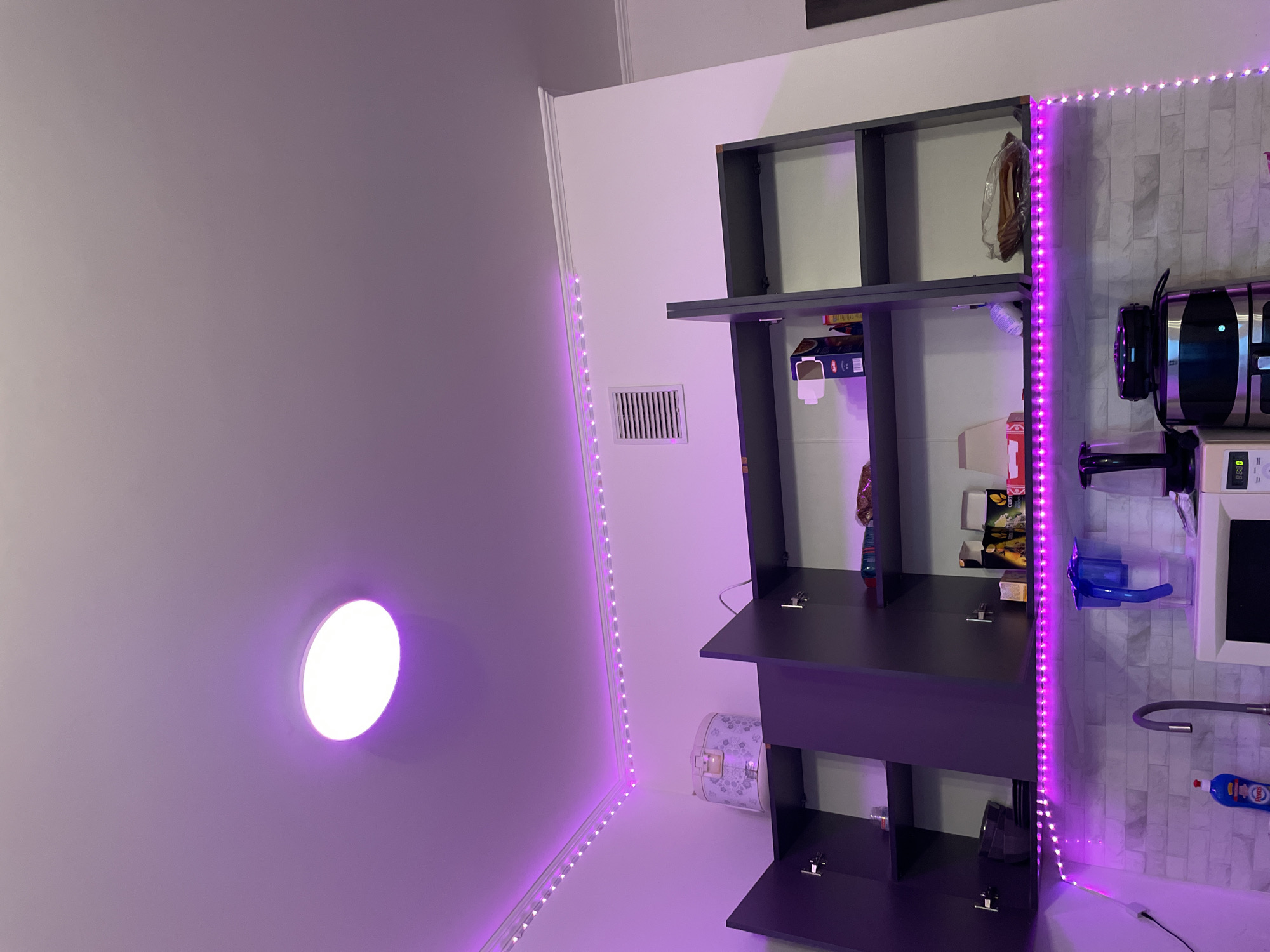 The image shows an indoor scene, likely a kitchen, with purple LED strip lighting illuminating the space. A round ceiling light is also visible, emitting a bright white light that is tinted purple by the ambient lighting. Above the cabinets, a decorative crown molding runs along the ceiling. On the top shelf of the cabinets, a white rice cooker with a floral pattern is visible. The cabinets themselves are dark gray and appear to be open, revealing various items stored inside, including boxes of food and what looks like a bag of bread. Below the cabinets, a countertop with a tiled backsplash features a kitchen sink with a modern faucet, a blue dish soap bottle, a water filter pitcher, and a coffee maker. A microwave is also visible on the counter. The overall impression is a modern and functional kitchen space enhanced by atmospheric lighting. The location of Ladozhskoe Ozero, Russia is not depicted in the image.