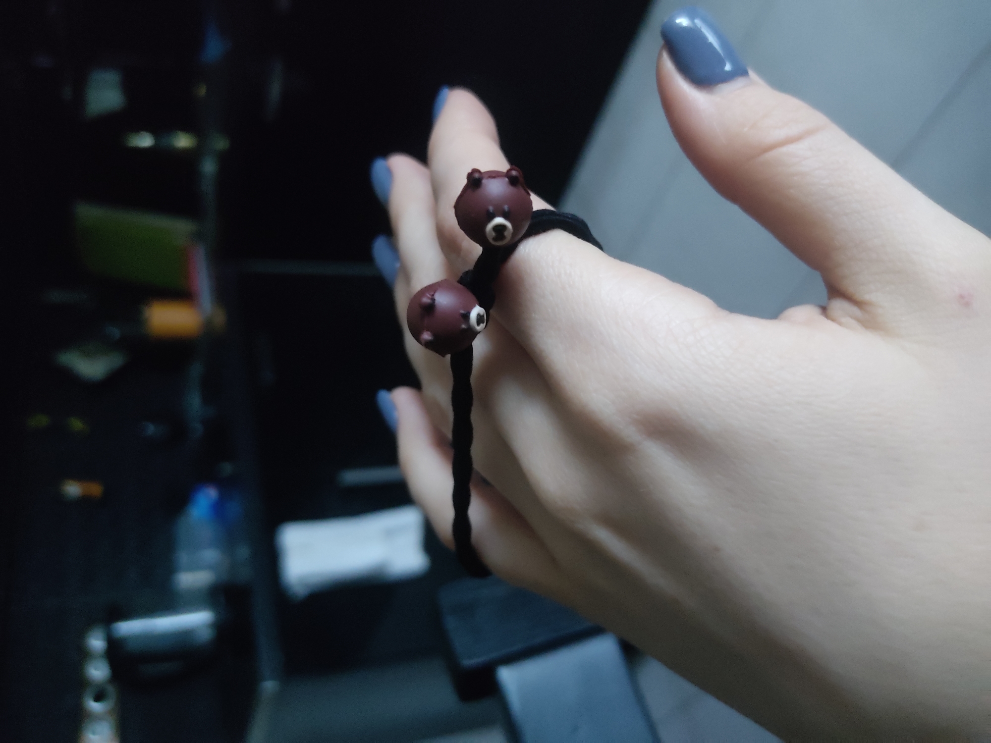 A right hand, adorned with grey-blue painted fingernails, holds a black elastic hair tie featuring two small, brown bear figurines. The background reveals a blurred and dark interior, potentially a vehicle, displaying a water bottle and various undefined objects. The location appears to be indoors, not identifiable within a city.
