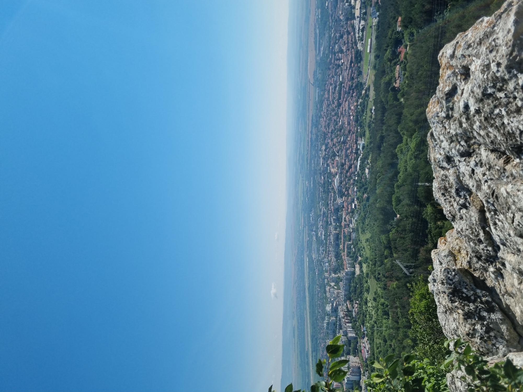 The image shows a panoramic view of the city of Gorna Oryahovitsa, Bulgaria, taken from an elevated viewpoint. In the foreground, there is a large, rough rock formation covered in lichen, with some green foliage visible on the left side. Beyond the rocks, the city sprawls across the landscape, characterized by a dense collection of buildings with red tile roofs and some taller, modern structures. A significant area of green forest separates the rocky foreground from the city, with a power line pylon visible within the trees. The middle ground features more of the urban landscape, interspersed with green spaces. In the background, a hazy horizon line suggests plains or rolling hills, with a faint strip of sky transitioning into a clear, bright blue sky overhead. The overall scene is one of a city nestled within a natural environment, viewed from a high vantage point on a sunny day.