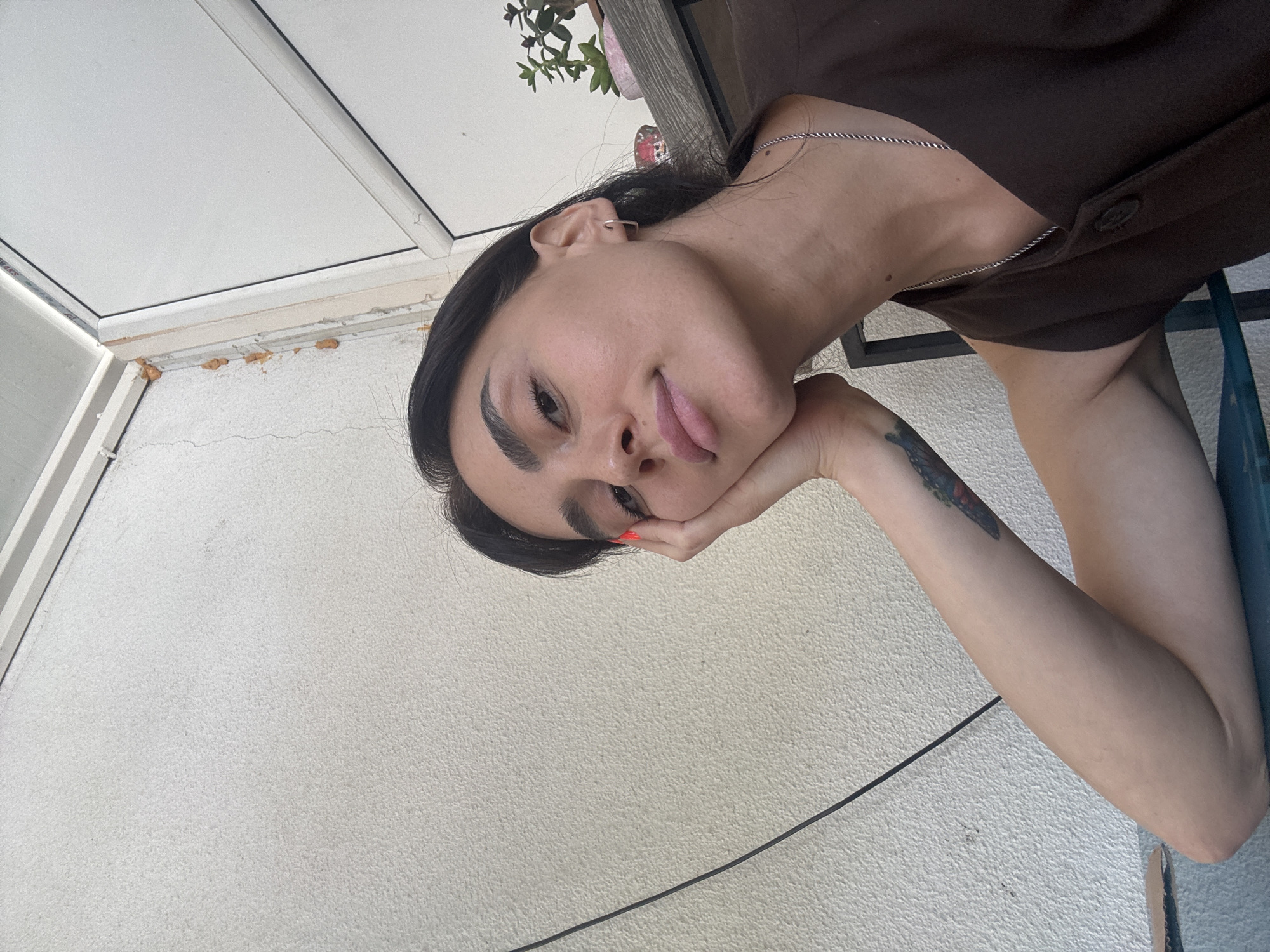 The image depicts a close-up, selfie-style shot of a young woman resting her cheek on her hand. She has dark hair, prominent eyebrows, and is looking towards the camera with a neutral expression. She is wearing a dark brown sleeveless top and a silver chain necklace. A tattoo is visible on her left forearm. The background appears to be an outdoor setting, with a textured light-colored wall and part of a white ceiling with visible sealant. To the right, a wooden shelf holds a small potted plant. A black cable runs diagonally across the left side of the frame. The lighting suggests it is daytime. No specific landmarks identifiable as being in Tbilisi, Georgia are present.
