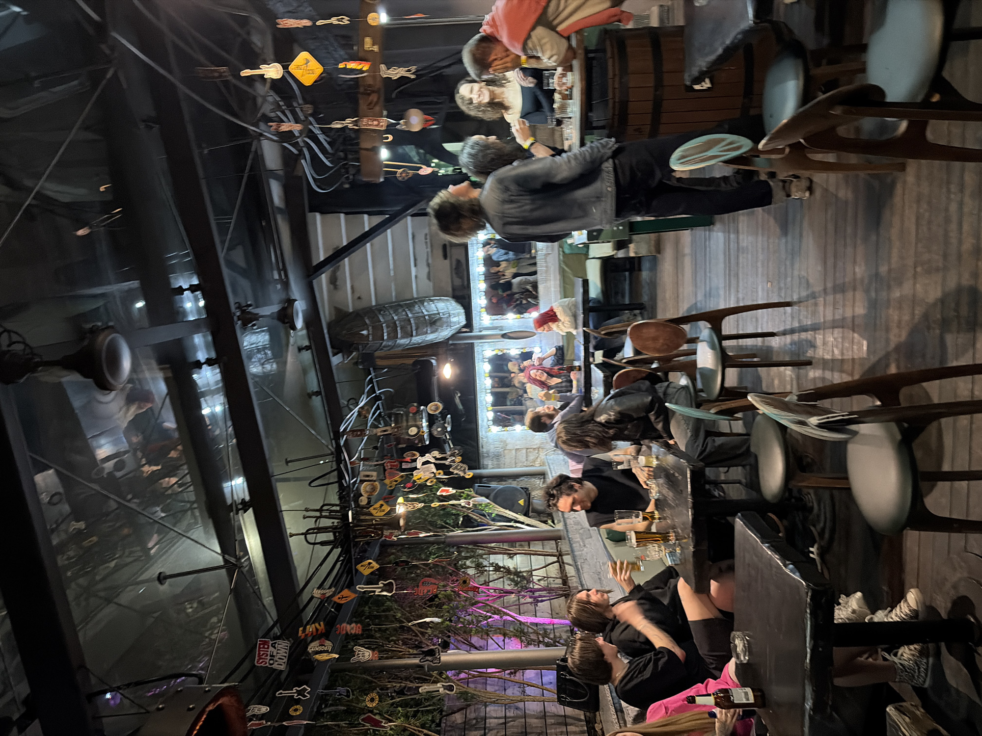 The image depicts an indoor bar or restaurant, likely in the city of Minsk, Belarus. Patrons are seated at tables, drinking and conversing. The establishment features a high ceiling with exposed structural steel and is decorated with hanging signs, guitar paraphernalia, and greenery. There's a barrel serving as a high table at one end, and other tables are arranged with chairs of varying designs. People are generally casually dressed and engaged in typical bar/restaurant activities.