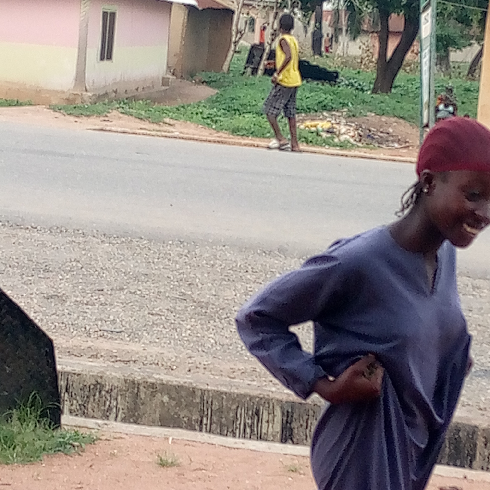 The image depicts a street scene in Mararaba, Nigeria. In the foreground, a woman with dark skin is wearing a burgundy head covering and a long-sleeved blue dress. She is smiling and walking, with her hand resting on her hip. In the background, a young man is walking on the side of the road. He is wearing a yellow shirt and checkered shorts. There is some scattered litter and debris along the roadside. Further in the background, there are trees and buildings. The road is grey with a concrete barrier. The sky is not visible.