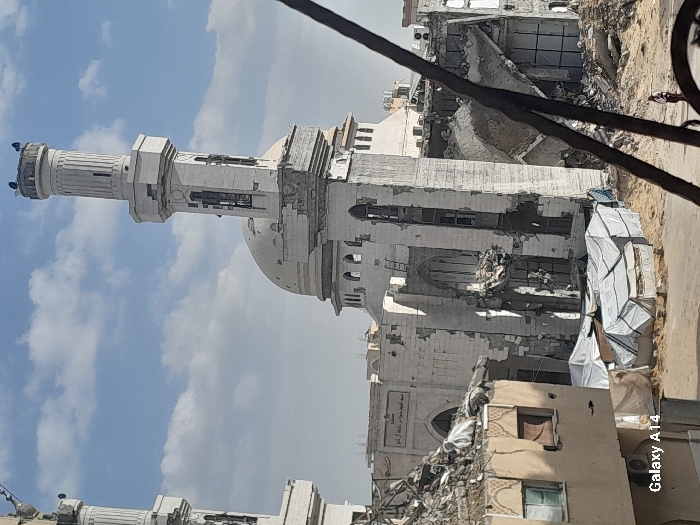The image displays the significantly damaged Mosque of Lady Aisha (مسجد السيدة عائشة أم المؤمنين) in Khan Yunis, Palestinian Territory, under a sky with scattered white clouds. The mosque's light-colored structure exhibits extensive destruction: its central dome has multiple visible impact points, and the prominent right minaret shows severe structural compromise with missing sections and fractures. The main building walls are largely destroyed, revealing internal rubble and broken arched window frames. In the immediate foreground to the left, a multi-story building is also heavily damaged, with its roof partially collapsed and debris on its upper level. Below the mosque and in the middle ground, several makeshift shelters constructed from white tarpaulins and other materials are erected on sandy, debris-laden ground. A single individual, dressed in light-colored clothing, walks on a dirt path in the lower right portion of the frame, moving away from the viewer. A dark, diagonal structural element, possibly a pole or cable, partially obstructs the right side of the scene.
