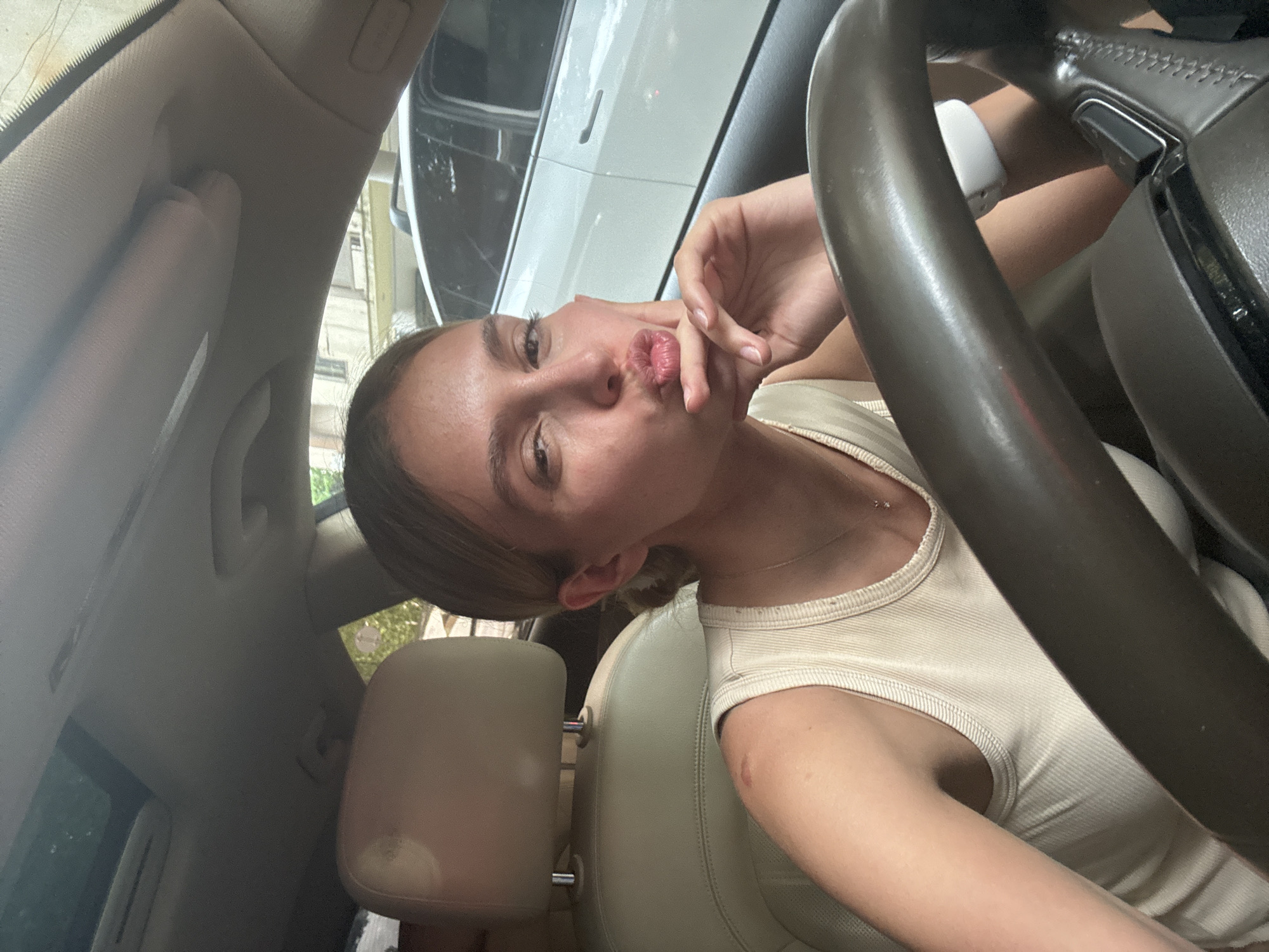 A young woman is in the driver's seat of a car. She has light brown hair and a light complexion. She is wearing a tan tank top and a white watch. She is pouting and touching her face. The car is parked, as evidenced by the position of the steering wheel. A white vehicle is visible through the car window. The location is undetermined.