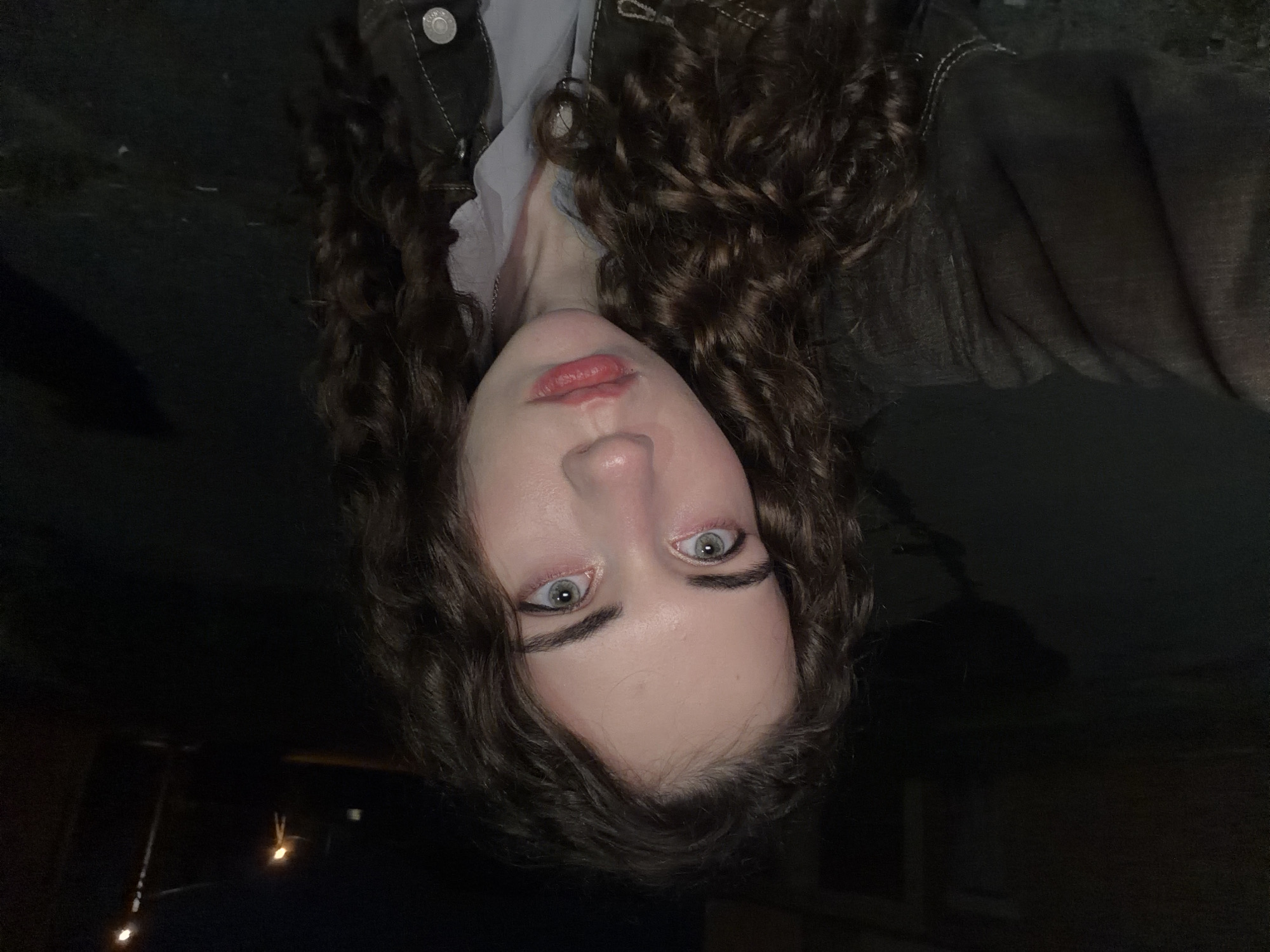 A nighttime, self-taken, eye-level portrait of a young woman with curly brown hair, pale skin, and light-colored eyes. She is wearing a brown jacket over a light-colored shirt. Her expression is neutral, with her lips slightly parted. The background is dark and appears to be an outdoor urban setting, possibly on a street or road, with a street light visible in the distance. The location is identified as Zavodoukovsk, Russia.
