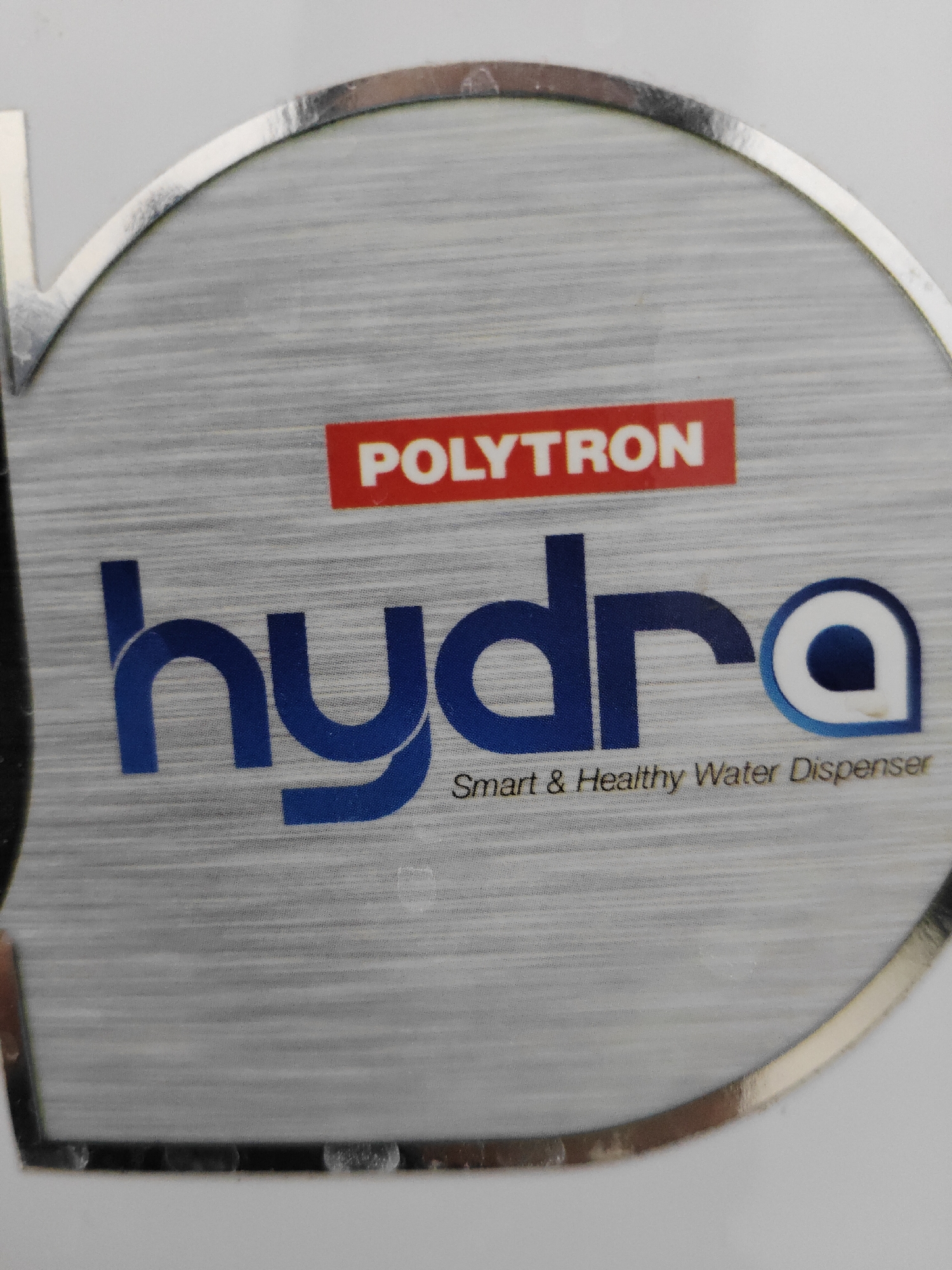This is a close-up, high-angle, eye-level shot of a label on a white appliance, likely a water dispenser. The label features a silver, brushed metallic background with a circular border. At the top of the circle, a red rectangular badge contains the word "POLYTRON" in white capital letters. Below this, the word "hydra" is prominently displayed in large, bold, blue sans-serif font. The letter "a" in "hydra" is stylized with a white circle outline containing a blue inner circle. Underneath "hydra," in smaller, light brown text, is the slogan "Smart & Healthy Water Dispenser." The lighting is bright and even, with no harsh shadows. The image appears to be taken in an indoor setting, possibly a home or commercial space. There are no people visible in the image. The location within the city of Semambung, Indonesia, is not discernible from the visual information provided in the image itself.