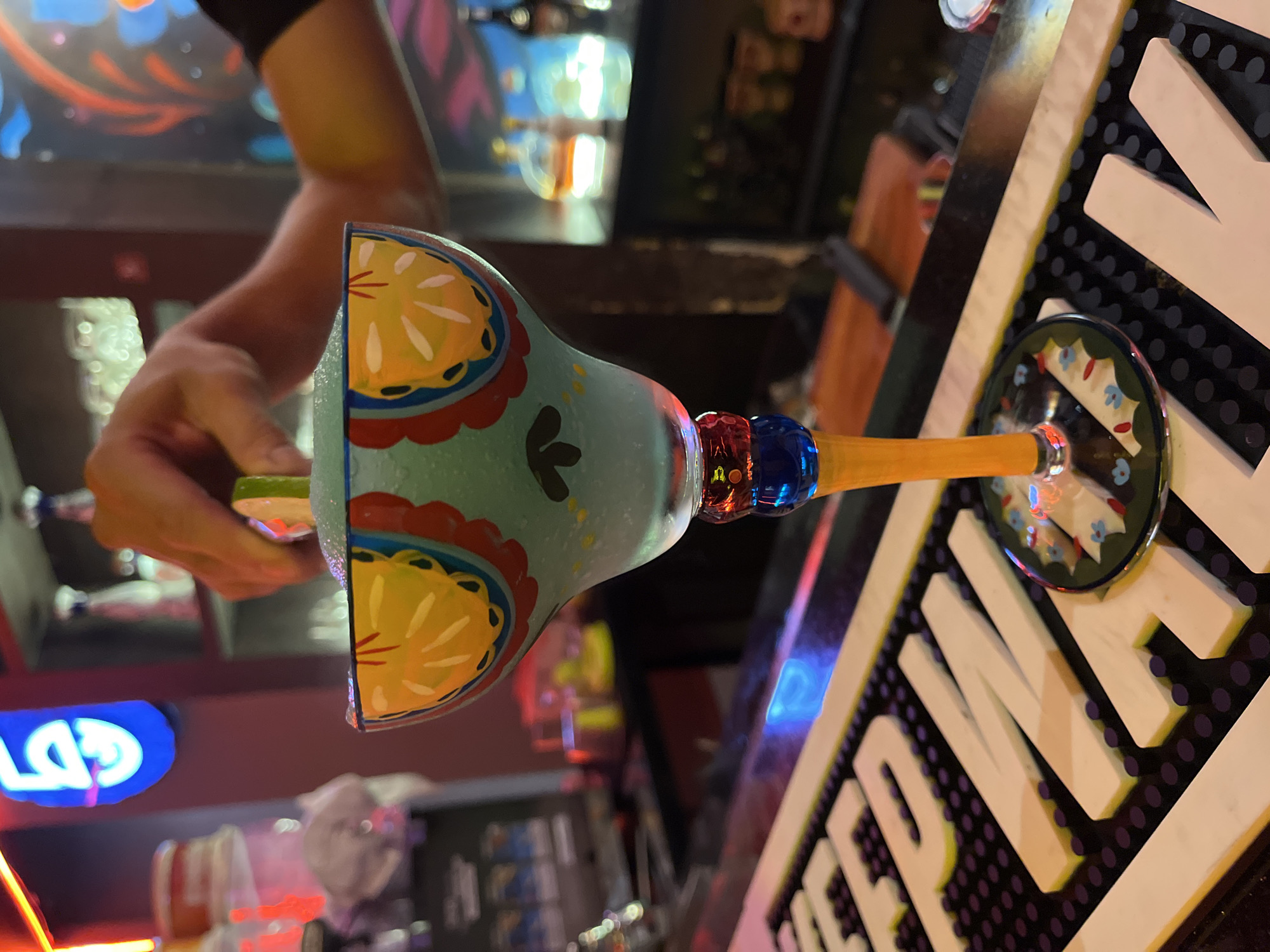 A person's hand holds a lime wedge near the rim of a brightly decorated margarita glass. The glass is turquoise with yellow and red designs. It has a long, orange stem and a decorated foot. The glass rests on a black and white bar surface. The background shows a bar setting, with illuminated signage and bottles visible. The location is inferred to be Đà Nẵng, Vietnam.