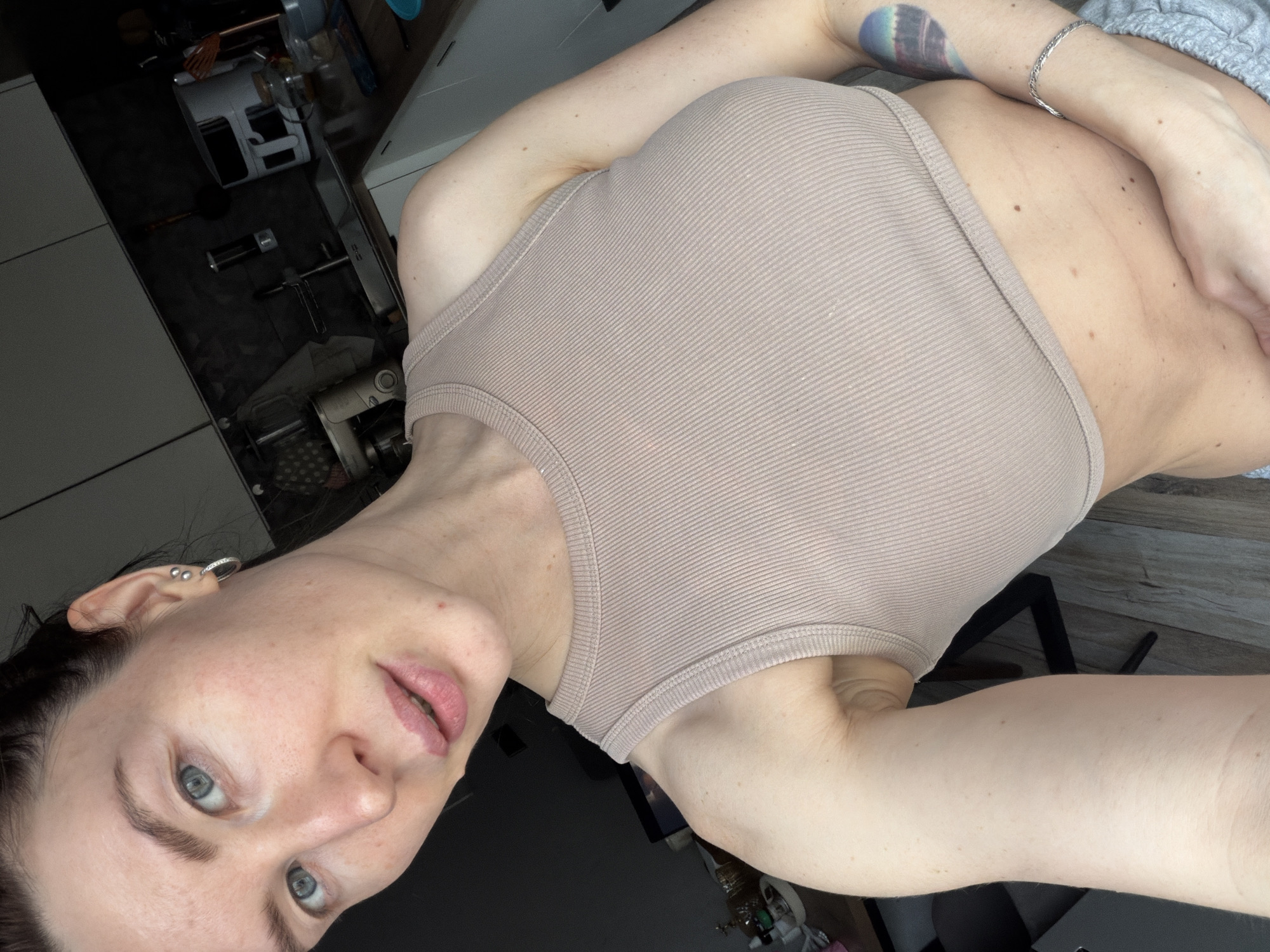 A person with fair skin, light-colored eyes, and dark hair is visible, wearing a light brown tank top and a silver bracelet on their left wrist. A tattoo is visible on the same arm. The person is positioned indoors, with kitchen appliances and countertops discernible in the background. The location is consistent with an urban environment, possibly Perm, Russia.