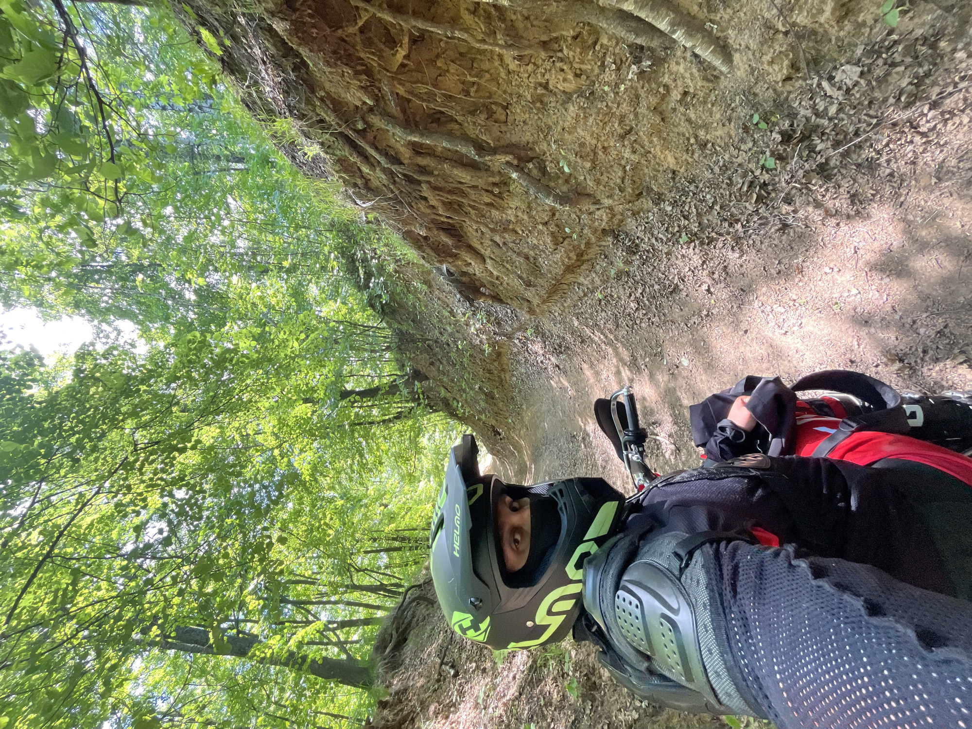 A person wearing a black and green helmet, mesh shirt, and body armor is on a motorcycle on a dirt path. The path is bordered on one side by a dirt wall with exposed roots, and on the other side by a forest with green trees. The location appears to be a rural area in Новочувашский, Russia.