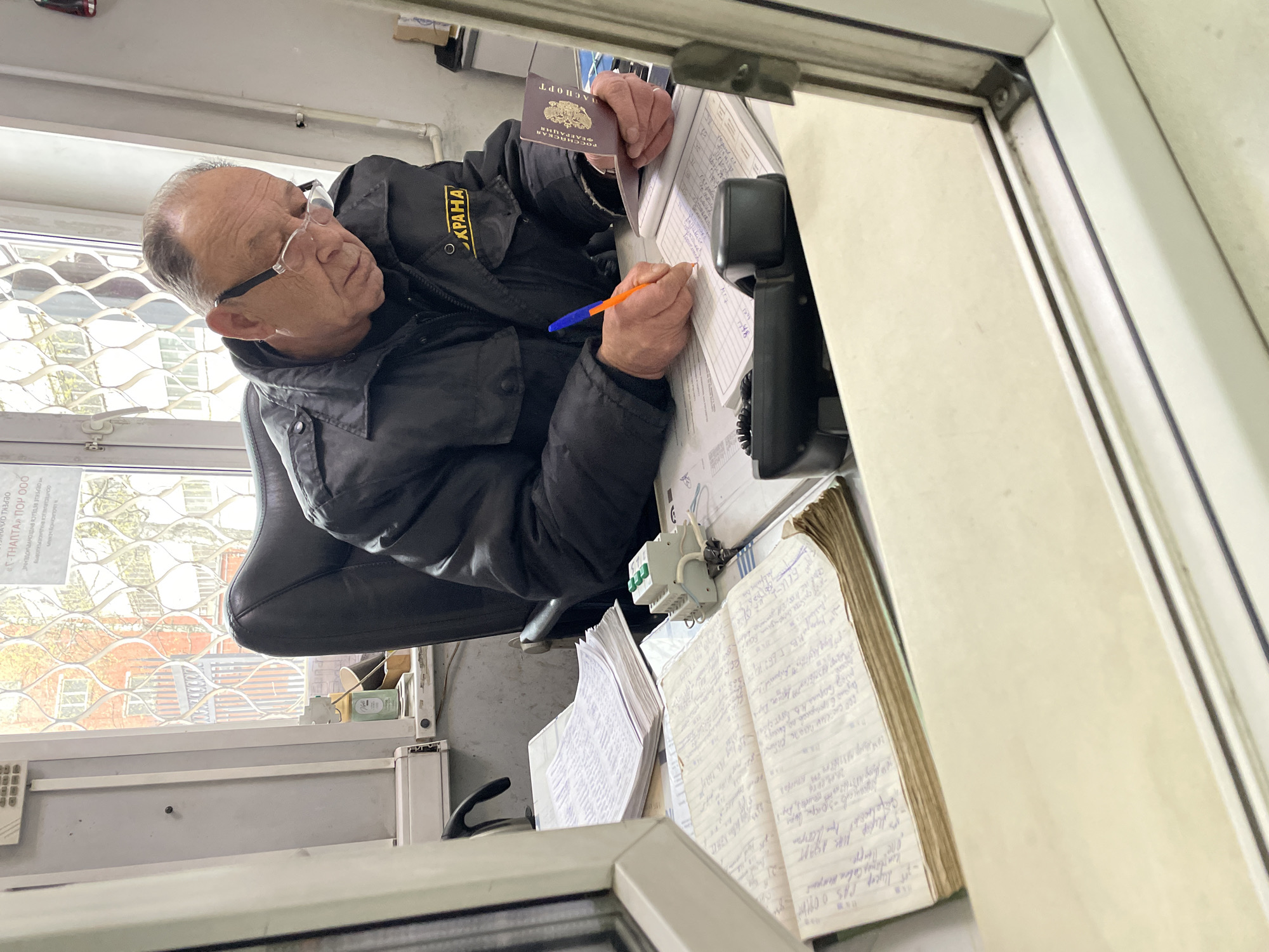 An older man wearing glasses and a black jacket with "ОХРАНА" (Security) written in yellow is seated at a desk, focused on writing in a notebook with an orange and blue pen. He holds a maroon Russian passport, clearly displaying "ПАСПОРТ" (Passport) and "РОССИЙСКАЯ ФЕДЕРАЦИЯ" (Russian Federation) on its cover.
The setting appears to be a security post or office, characterized by a functional, somewhat cluttered workspace. On the desk, there are multiple notebooks, stacks of papers, a black telephone, and a white electrical terminal block. A window with a security grate is visible behind the man, through which an urban or industrial background can be vaguely seen. A sign on the window also bears the text "«Γ-ТНАПТА» ПОР ООО", likely indicating a company or organization, and "ОХРАНА". The man's expression is one of concentration as he performs his task, possibly logging entries or processing information.