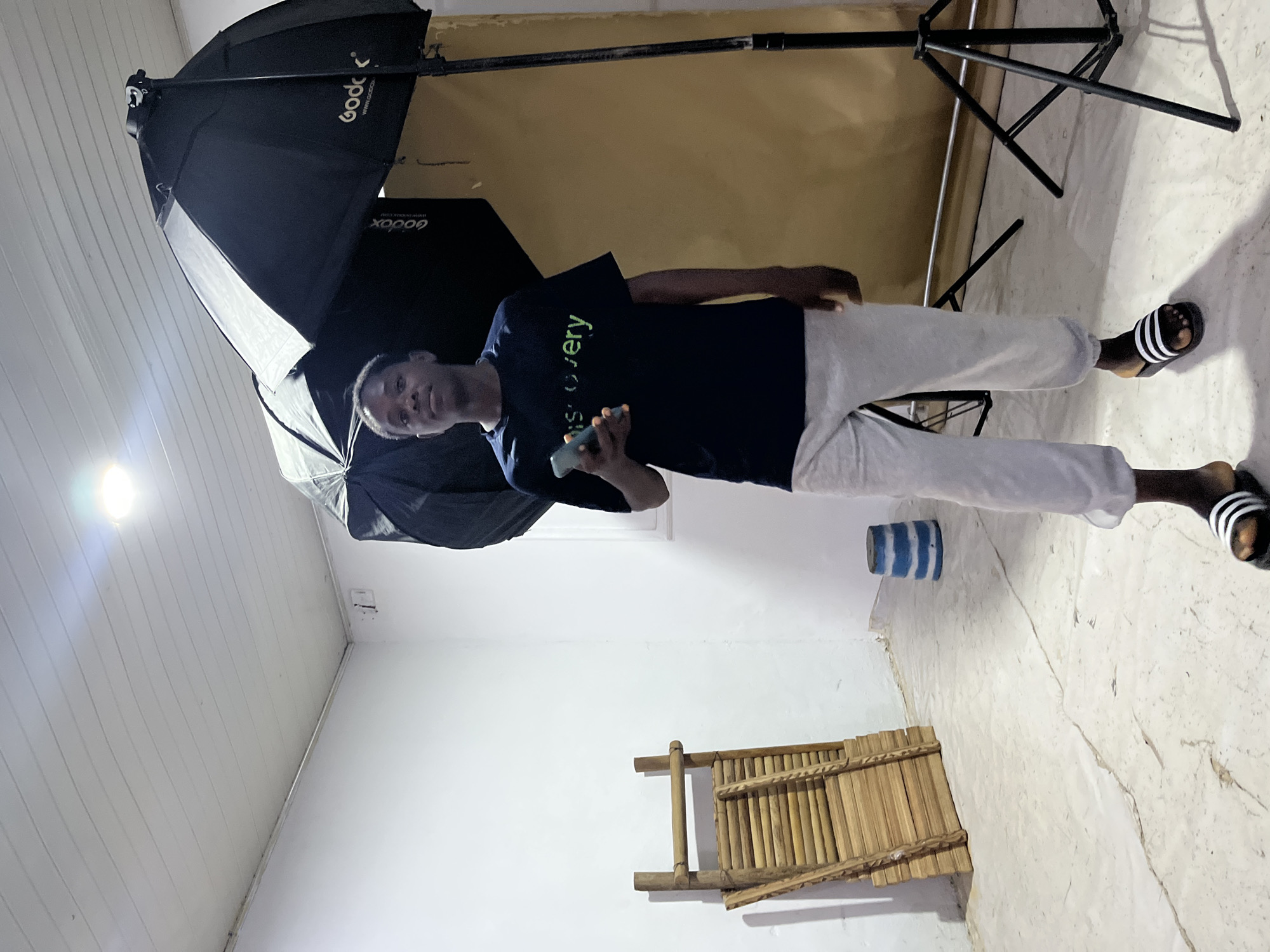 A person is standing indoors in a room with a white ceiling, a white wall, and a wall that appears to be painted beige. The individual is holding a cell phone. A bamboo flute is mounted on the wall, and a blue and white striped object sits near the floor. A photography lighting umbrella with the brand name "Godox" is visible, along with a lighting stand. The person is wearing a dark blue t-shirt with the word "discovery" on it, light gray pants, and black and white striped sandals.