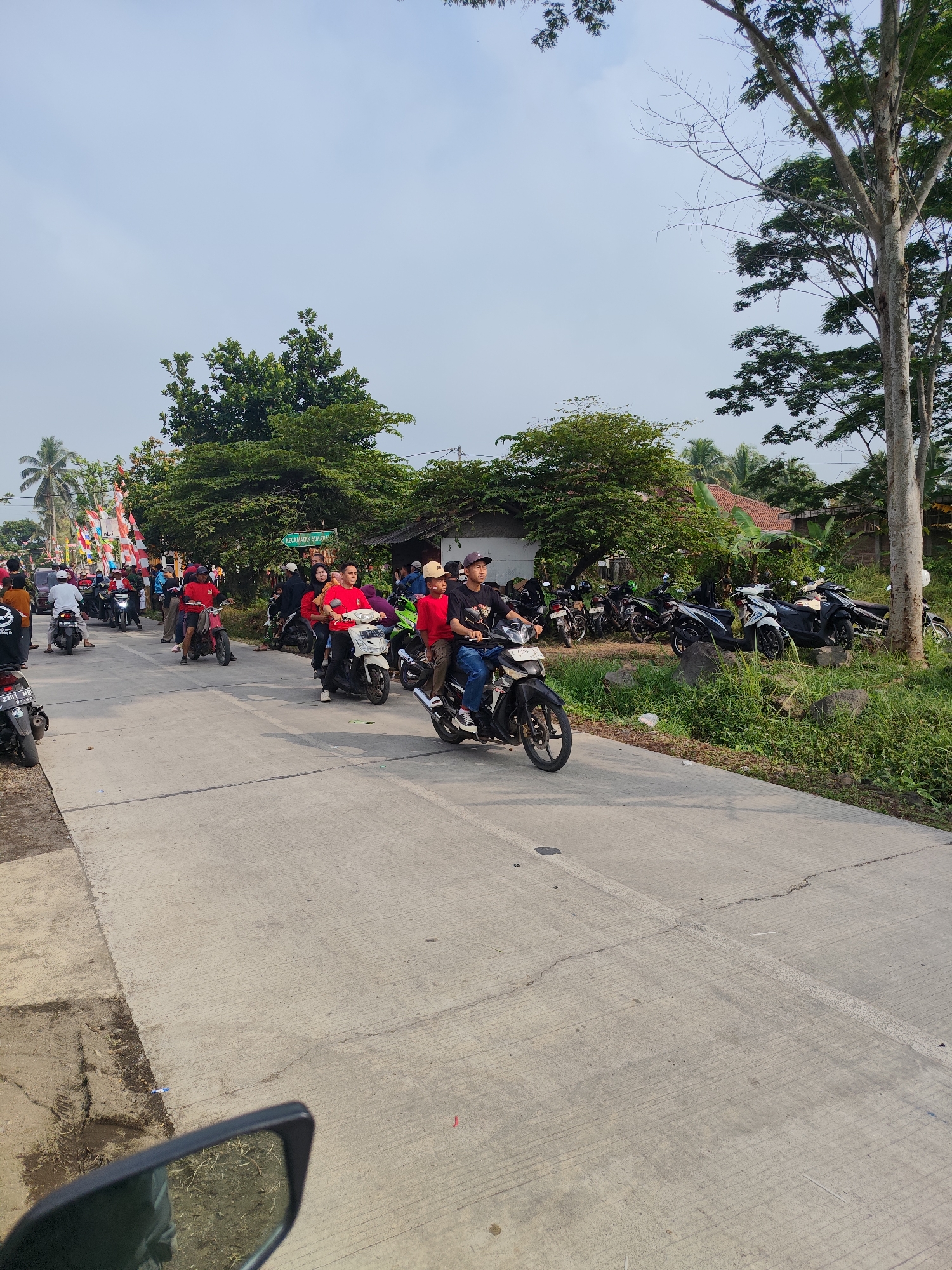 The image depicts a street scene in Sukamahi, Indonesia, likely during a festive occasion given the numerous motorcycles, red attire worn by many individuals, and Indonesian flags decorating the roadside. A procession of motorcycles, some with multiple riders, is traveling along a paved road. Several individuals are riding motorcycles, with some appearing to be part of a parade or convoy. Others are standing by the roadside, observing the event. In the background, there are trees, houses, and a street sign indicating "KECAMATAN SUKAMA..." suggesting the location is within Sukamahi district. The weather appears to be clear, with a bright sky. The overall atmosphere suggests a community event or celebration.