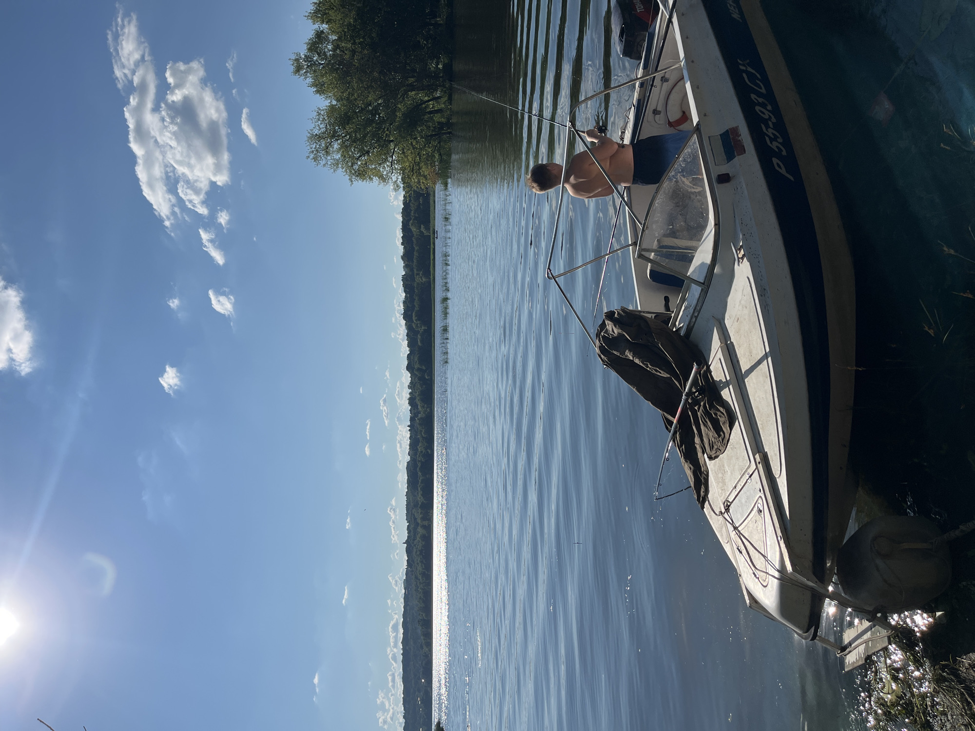 This image depicts a serene summer day on the Volga River in Saratov, Russia. The sun shines brightly in a clear blue sky dotted with fluffy white clouds. The water is calm, reflecting the sunlight and the distant treeline of the Saratov Hills.
In the foreground, a small white motorboat is moored near the shore. A young boy, shirtless and wearing blue swim trunks, stands at the back of the boat, fishing with a rod. The boat has a blue stripe along its side and a license plate that reads "P55-93CX". A life preserver is visible inside the boat, and a dark green tarp is draped over the front. The boat appears to be tied to the shore with a rope and a large, round fender. The shoreline is covered with sparse vegetation and debris.
In the distance, the city of Saratov is visible along the opposite bank, with buildings and trees forming a picturesque backdrop. The overall impression is one of peaceful recreation and enjoyment of nature on a warm summer day.