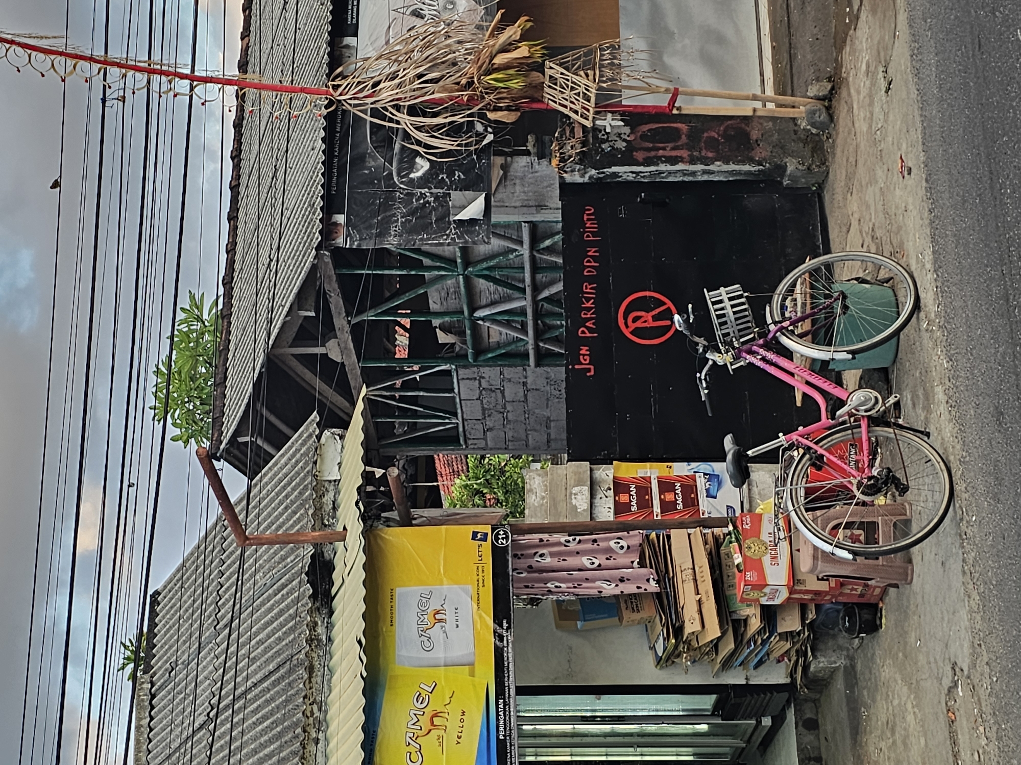 The image depicts a street scene in Denpasar, Indonesia, likely in an urban or semi-urban area, with a focus on a pink bicycle parked in front of a dark-colored gate. The gate has a red circular "R" symbol with a diagonal line through it, indicating "No Parking," and text in red that reads "Jgn Parkir Dpn Pintu," translating to "Don't Park In Front of the Door." To the left of the gate, there are stacks of cardboard boxes and some red boxes of what appear to be beverages, along with a pink curtain with a panda pattern hanging from an awning. Above the awning is a large yellow banner advertising Camel cigarettes, featuring images of yellow and white cigarette packs and text such as "SMOOTH TASTE" and "SINCE 1913."
The background reveals more of the urban environment. Several buildings with corrugated metal roofs are visible, some with visible wooden structural elements. Overhead, a complex network of electrical wires crisscrosses the sky. The sky itself is overcast with hints of daylight. A small bird is perched on one of the wires. To the right of the gate, a red decorative pole with dried foliage and small woven structures is positioned near the roadside. The pavement in front of the buildings is concrete, showing some wear and tear, with debris scattered on the ground. The overall impression is one of a functional, perhaps somewhat cluttered, commercial or residential street in Denpasar.