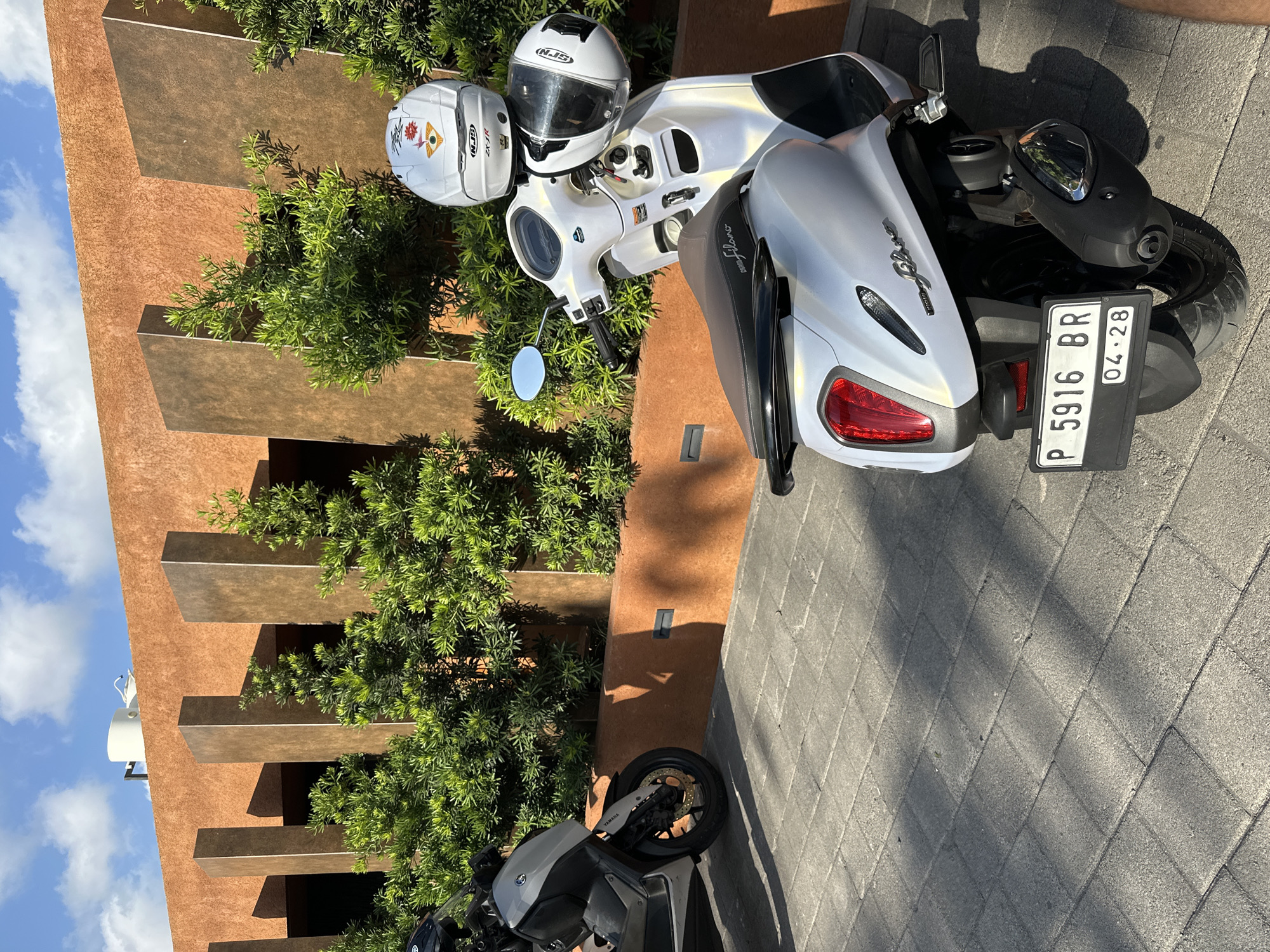 In Kuta Selatan, Indonesia, two parked scooters are visible on a paved pathway. The scooter in the foreground is white and pearlized, with a brown seat, and features two white helmets stacked on its handlebars. The rear of this scooter displays a license plate reading "P 5916 BR 04.28". To its left, a silver and black scooter, identified by a "YAMAHA" logo on its side, is partially visible. The scooters are situated in front of a building with a textured orange facade, punctuated by tall, dark rectangular pillars and lush green shrubbery. The sky above is a clear blue with scattered white clouds, and sunlight casts shadows on the pathway.