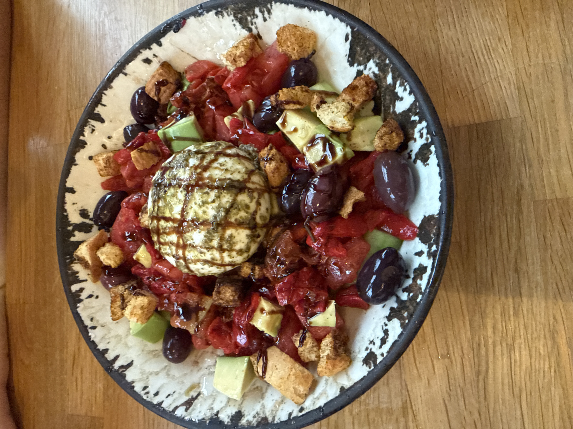 A high-angle shot depicts a ceramic bowl of food, placed on a light brown wooden surface. The bowl has a speckled pattern of white and black around the rim and interior. The food consists of a large, spherical, off-white ball, drizzled with a dark, crisscrossing sauce. Surrounding the ball are chunks of red tomatoes, diced avocado, dark olives, and small, golden-brown croutons. A similar dark sauce is drizzled over the salad. The background is a plain, light brown wooden surface. The location is not discernible from the image.