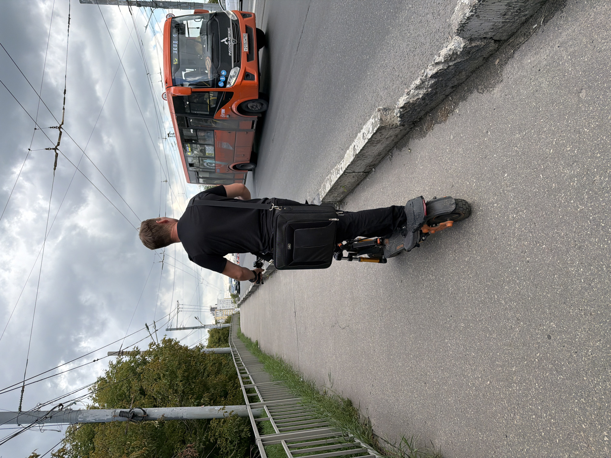 An adult male, viewed from behind, rides an electric scooter along a paved sidewalk in Nizhny Novgorod, Russia. The man is wearing a black t-shirt and black pants and has a black bag slung over his shoulder. He is moving away from the camera and to the right. Beside the sidewalk, a metal fence separates it from a grassy area and trees. Across the street, an orange bus is visible, with the words "РУССКИЙ АВТОБУС" printed on its front. The sky above is overcast with clouds. The scene appears to be a typical urban environment.