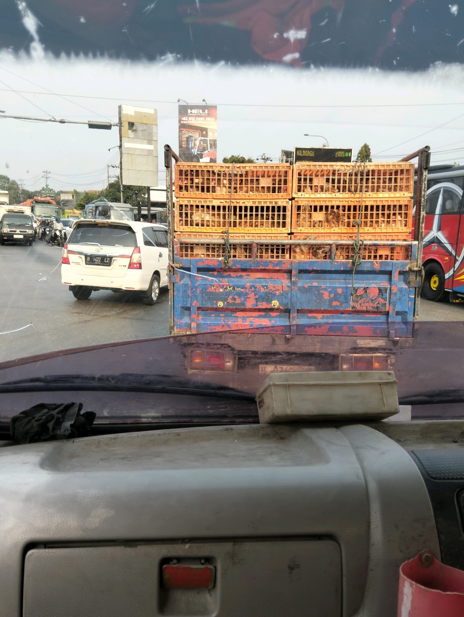 The image shows a street scene in Semarang, Indonesia, from the perspective of a vehicle. In the foreground, the dashboard of the vehicle is visible, with a dark cloth placed on the left side and a light-colored plastic container on top of the dashboard. In the midground, a blue truck is loaded with stacks of orange cages filled with chickens. The truck has some signs and graffiti on its side, including a picture of a man and text. To the left of the truck, a white car with license plate H 1422 LZ is visible, along with other vehicles and motorcycles on the road. In the background, there are buildings, power lines, and billboards, including one advertising "HELI". The sky is overcast with some white streaks. The overall impression is of a busy street in an urban environment.