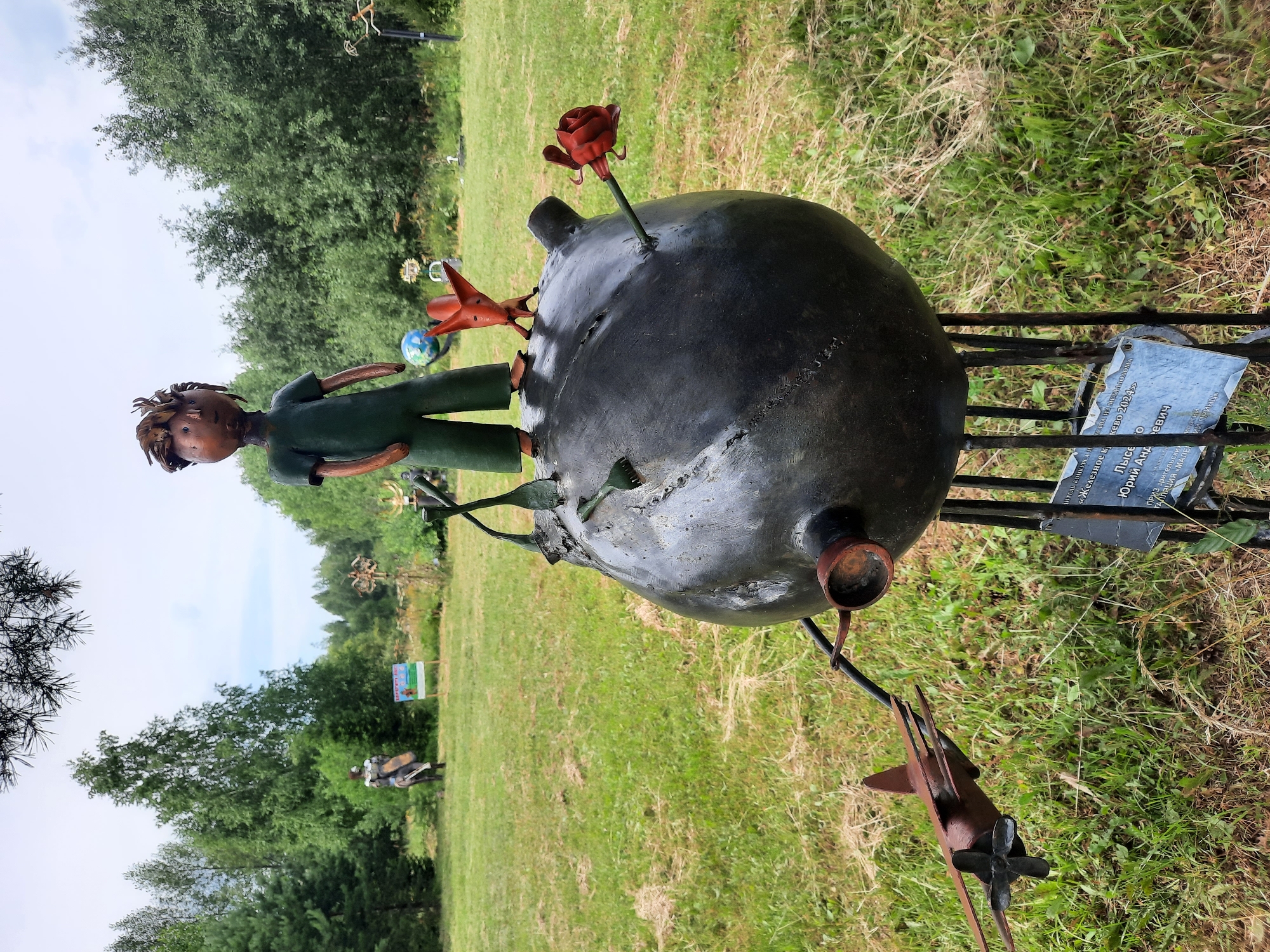 In the city of Zelenogorsk, Russia, a metal sculpture depicts the Little Prince standing atop a large, dark, spherical object supported by metal legs. The Little Prince, rendered in a simplistic style, wears a green outfit. Beside him on the sphere, a small red fox figure stands, and a blue globe is visible. A red metal rose is attached to the sphere. To the left of the sphere, a small, rusty metal airplane with a propeller is attached to an extending arm. A dark metal plaque is affixed to the sculpture's legs. In the background, a grassy field is dotted with trees under a cloudy sky. Further in the background, other sculptures and a signpost are visible.