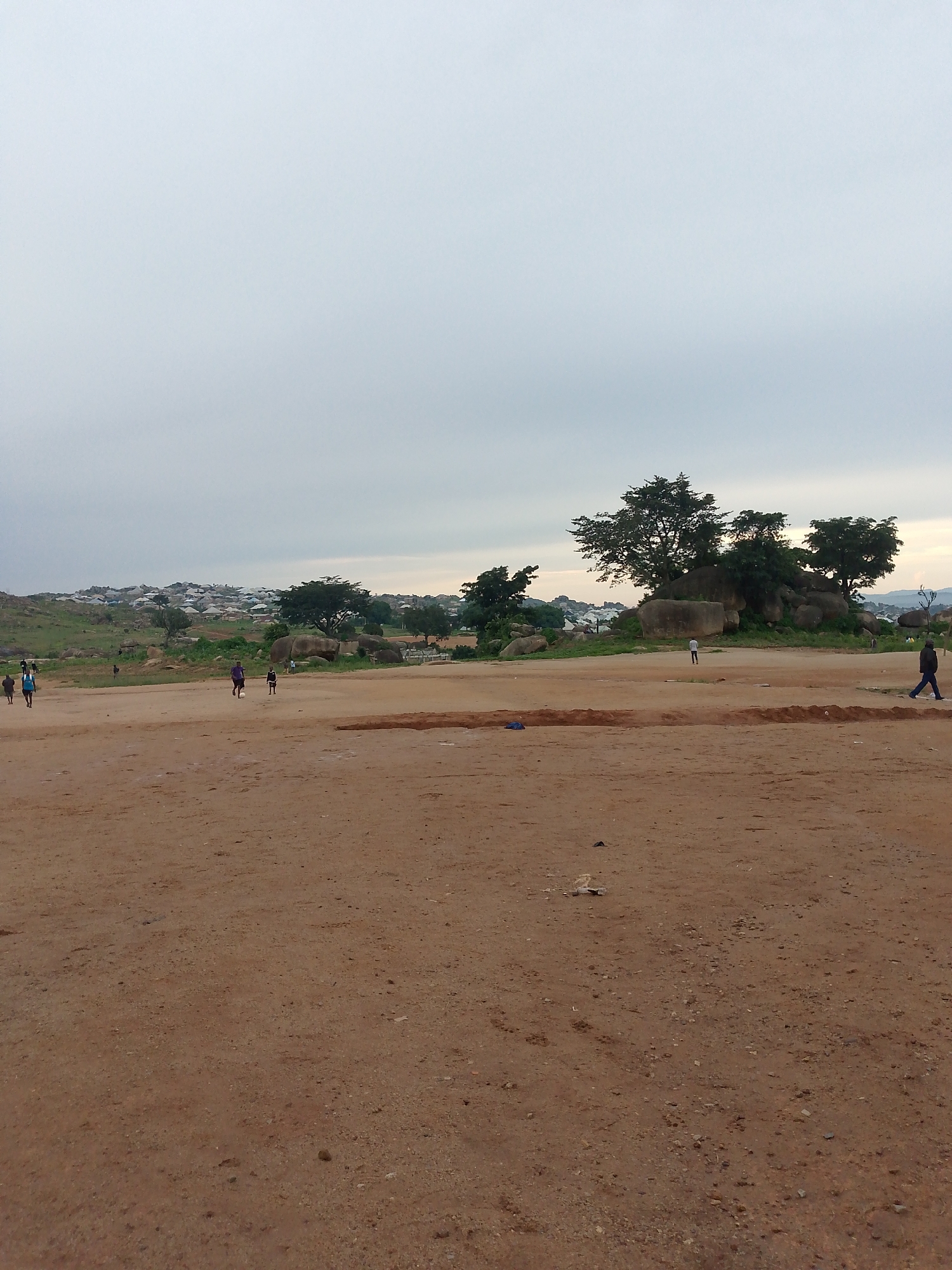 A wide-angle shot captures a vast, open dirt field with a few scattered individuals. In the foreground, the ground is a reddish-brown dirt, appearing dry and uneven with small stones. A trench or excavated area is visible in the middle ground, dividing the field. In the mid-distance, several people are walking or standing on the field. To the left, three figures are walking from left to right. Further into the field, a person in a purple shirt is walking towards the right, and another person in a dark shirt is walking to the left. In the background, a cluster of trees and large rocks are present, with a hillside populated by numerous small dwellings and buildings forming a settlement on the horizon. The sky is overcast with a pale, diffused light. To the far right, a person in dark clothing is walking away from the viewer.
