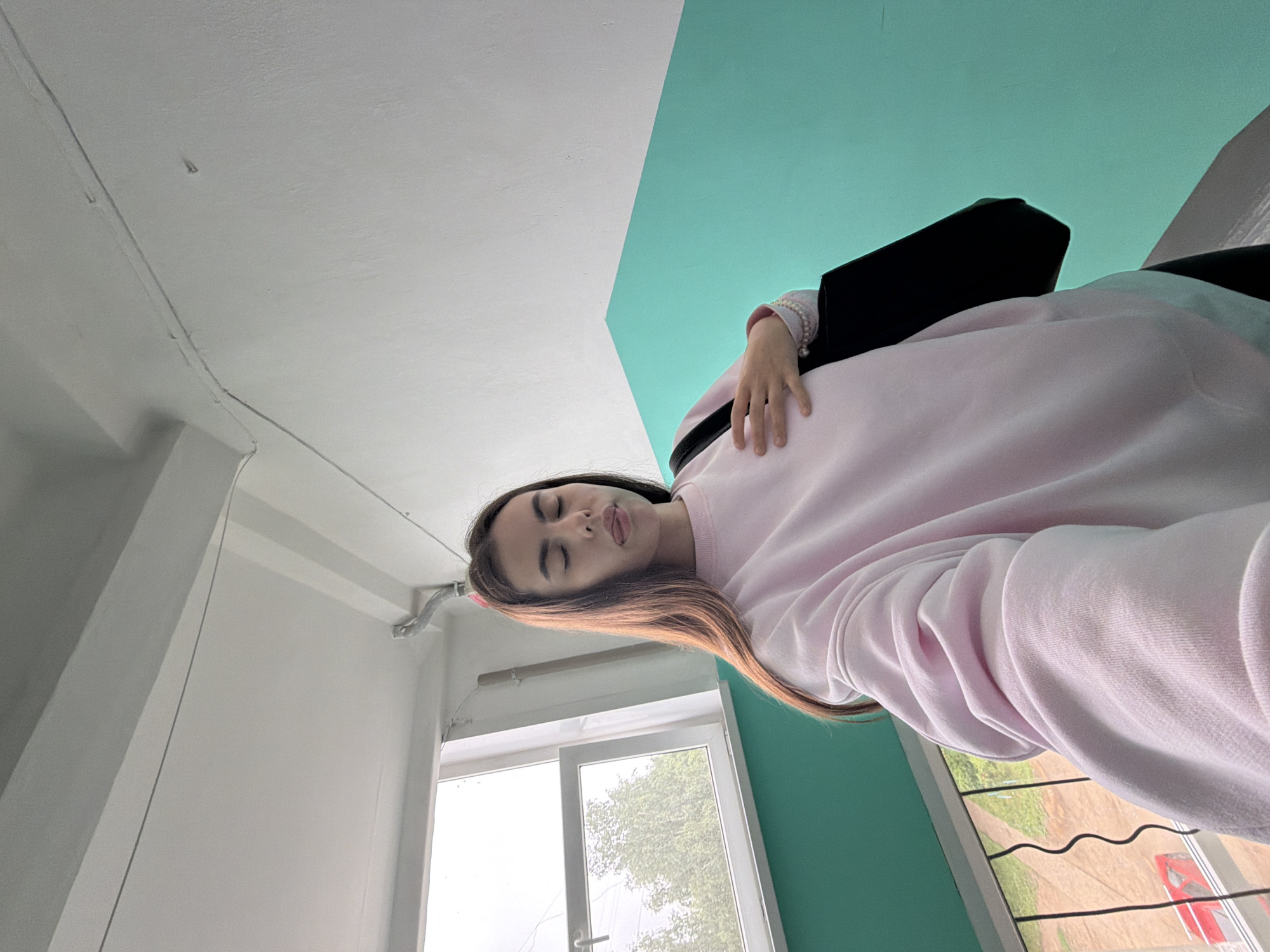 A low-angle indoor portrait features a young woman looking down towards the camera. She has long, dark brown hair with lighter ends and is wearing a light pink, long-sleeved sweatshirt. Her eyes are closed, and her tongue is slightly out, suggesting a playful or relaxed demeanor. On her left wrist, she wears a pearl bracelet, and her left hand rests near a black bag or object tucked under her arm.
The background consists of an interior space with a striking two-toned wall, split diagonally. The upper portion is white, while the lower section is a solid teal color. Exposed white wires or cables run along the white wall and ceiling. A white-framed window in the lower-left shows an outdoor scene, featuring green tree foliage and what appears to be a paved area or road with a red vehicle partially visible in the distance. The lighting suggests it is daytime, with soft natural light entering through the window. The composition is taken from below, looking up, providing a unique perspective of the room. There is no visible text in the image.