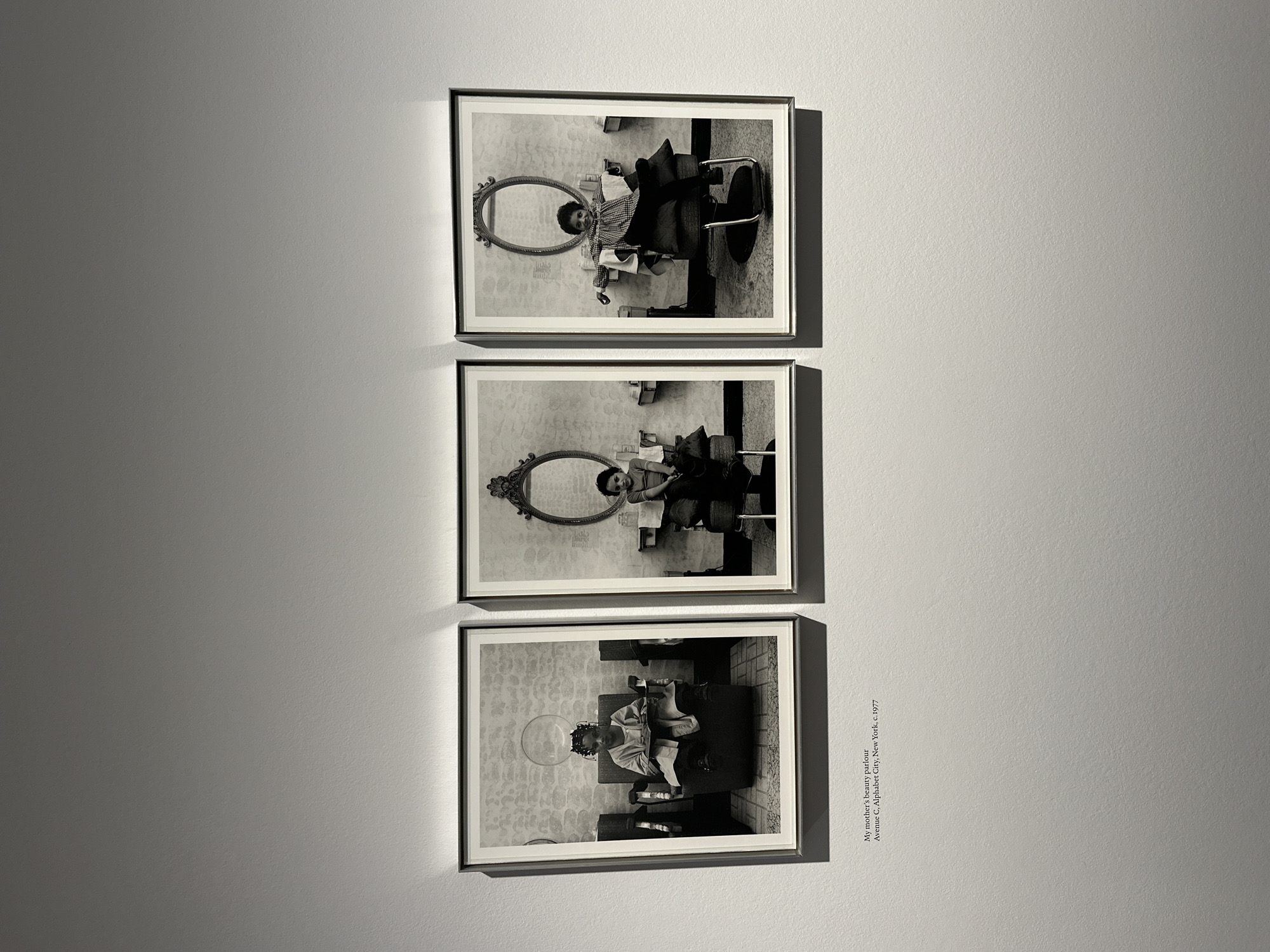 Three black and white framed photographs are displayed vertically on a plain, light grey wall in what appears to be an exhibition or gallery space. The overall image is taken in London, United Kingdom.
Each photograph captures a scene within a beauty parlor. In the top two frames, a person, possibly a woman or girl, is seen reflected in a large, ornate oval mirror, seated in a salon chair. The reflection shows the person looking towards the viewer. The background within these photos features patterned wallpaper. In the bottom photograph, a person is depicted from a slightly elevated, side-rear perspective, sitting in a salon chair with their hair under a hooded dryer.
The lighting in the room where the photographs are displayed creates shadows cast by the frames onto the wall, suggesting illumination from above.
Adjacent to the bottom framed photograph, text is printed directly onto the wall. It reads: "My mother's beauty parlour / Avenue C, Alphabet City, New York, c. 1977". This text likely serves as the title and descriptive information for the photographic series. The series documents scenes from a beauty parlor located in Alphabet City, New York, around 1977.