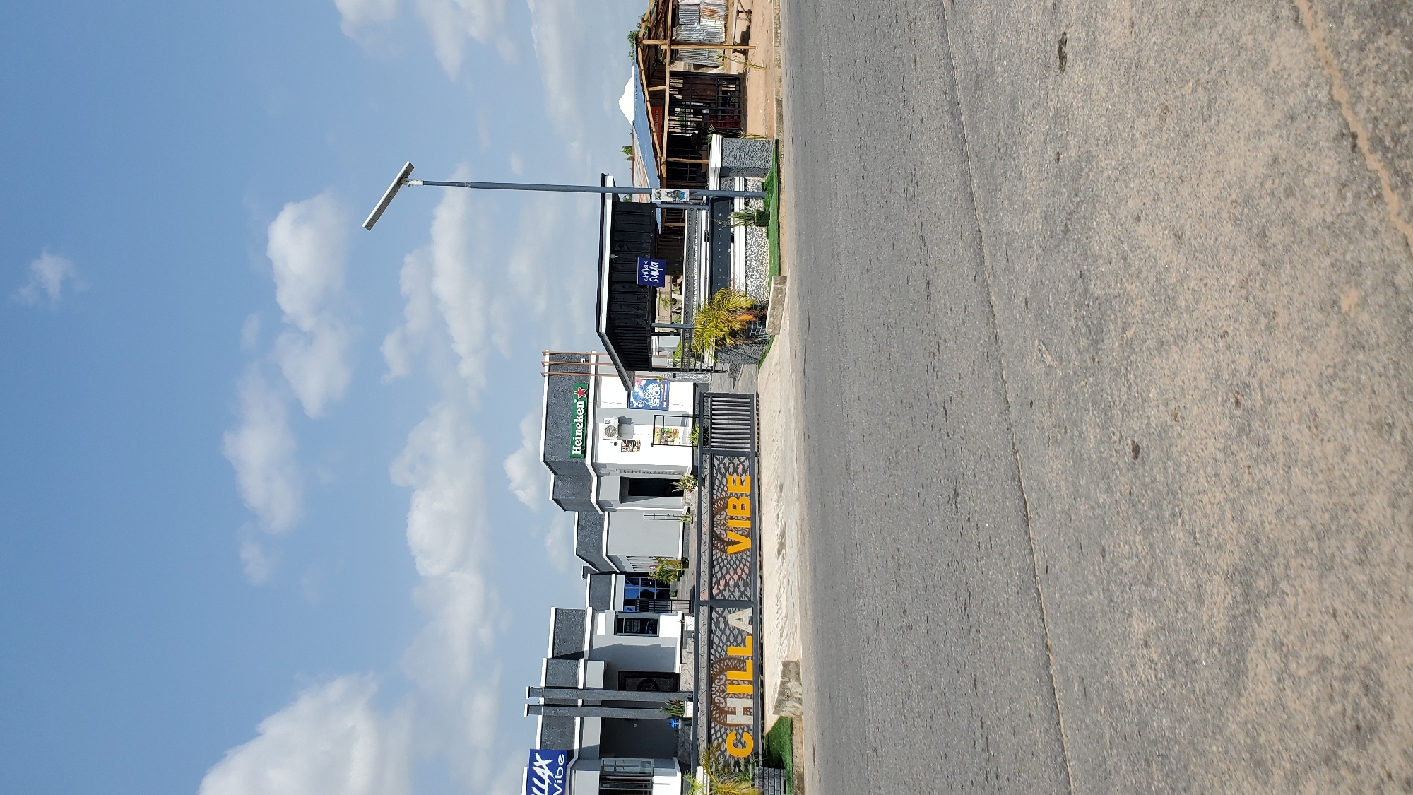 The image depicts the "CHILLA VIBE" establishment in Jalingo, Nigeria. The establishment features a modern architectural design with grey and white buildings. A prominent sign with "CHILLA VIBE" in yellow lettering is visible at the entrance, which is a gated area. The establishment has a Heineken advertisement displayed. Adjacent to the main building, there is an open-air structure with a thatched roof and wooden seating. The scene is set against a backdrop of a blue sky with scattered white clouds. The foreground shows a paved road. There are no people visible in the image.