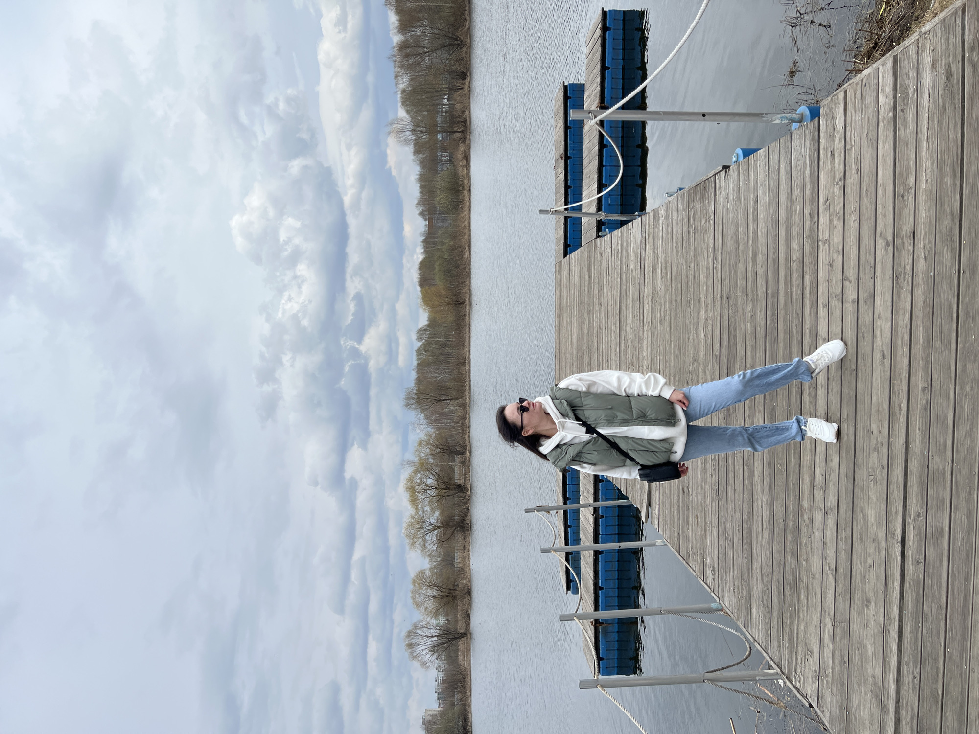 The image shows a woman standing on a wooden dock at an angle to the camera. The location is likely in the city of Kazan, Russia, based on the surrounding landscape.
The woman is wearing a light green and white puffy jacket, light blue jeans, and white sneakers. She has sunglasses on and a small black bag across her body. She is looking towards the sky, which is cloudy.
The dock is made of wooden planks and has blue plastic flotation devices underneath. There is water on either side of the dock, and trees line the opposite shore. Metal railings are visible on the dock next to the flotation devices.