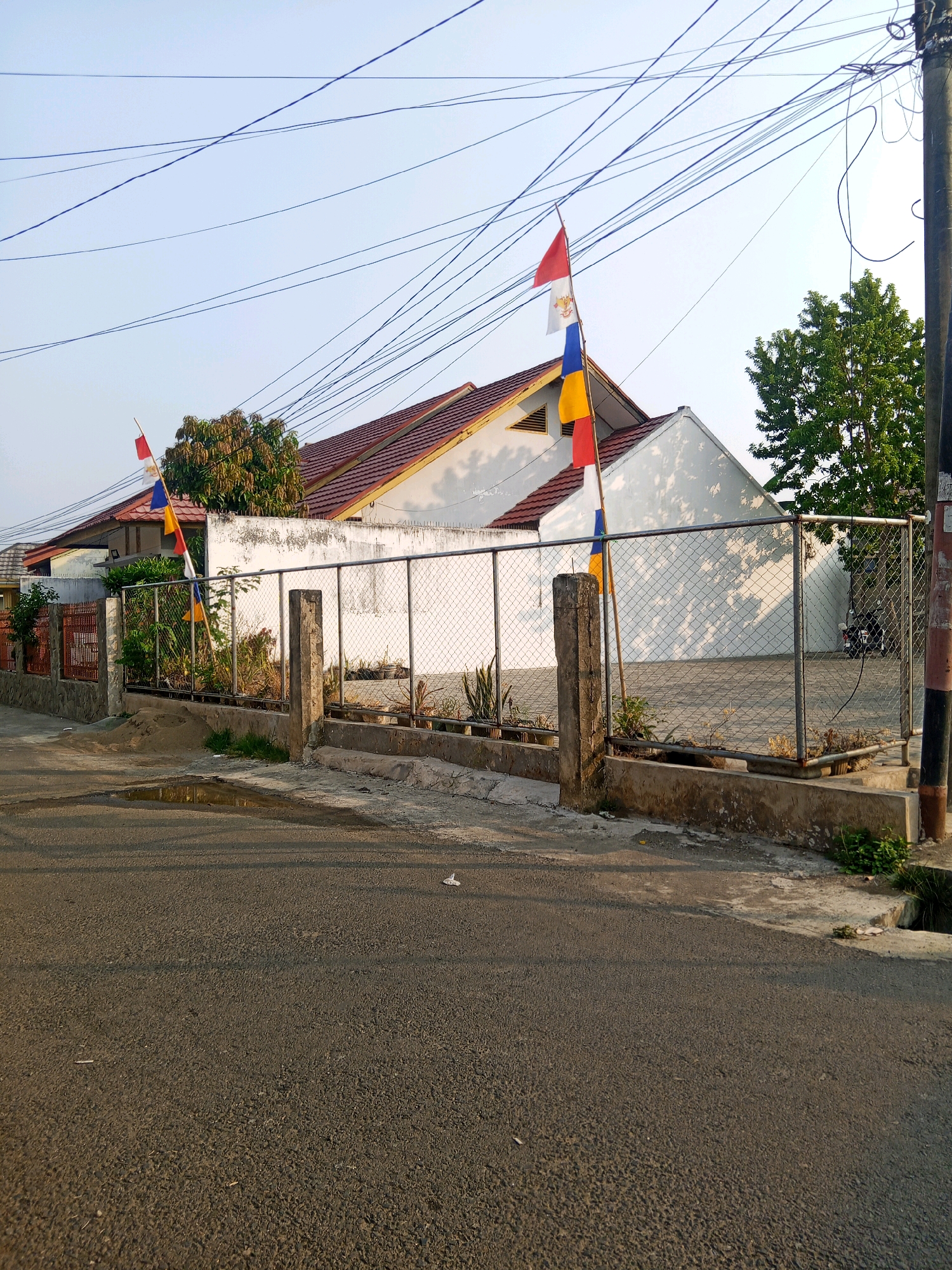 The image depicts a residential street scene in Lubuklinggau, Indonesia. In the foreground, a paved road with a puddle of water reflects the sky. A low concrete curb separates the road from a property, which is enclosed by a metal chain-link fence supported by concrete posts. Several Indonesian flags, in red and white, and other colors, are planted along the fence line. Beyond the fence, a white building with a red-tiled roof is visible, partially obscured by trees and foliage. The sky above is clear and a pale blue, with numerous electrical wires crisscrossing the frame. A tall utility pole is situated on the right side of the image. The overall impression is that of a quiet, sunlit residential area.