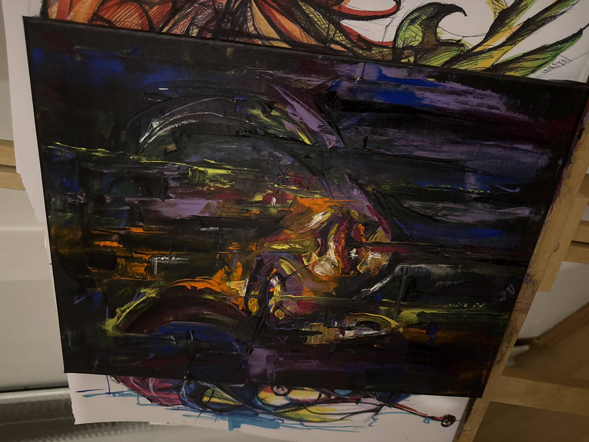 A close-up shot captures an abstract oil painting in progress, resting on an easel. The painting features bold brushstrokes of dark blue, purple, yellow, orange, and black. The artwork seems to depict a dimly lit scene, with the warmer colors clustered towards the lower center, suggesting a source of light or fire. The background is predominantly dark. The painting is placed on top of several drawings, which adds to the ambiance. The scene appears to be a studio, possibly in a location somewhere else rather than Sofia, Bulgaria, where paintings and drawings are created and stored.