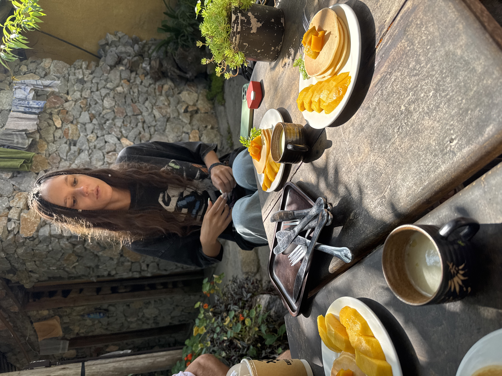 A woman with long brown hair sits at a wooden table in an outdoor setting in Lũng Cú Commune, Vietnam. She is wearing a black shirt and jeans. On the table are plates of pancakes and mango, along with cups and cutlery. A tray of cutlery rests in the middle of the table. There's a small potted plant in the background, and a stone wall is partially visible behind the woman. The sunlight casts strong shadows across the table.