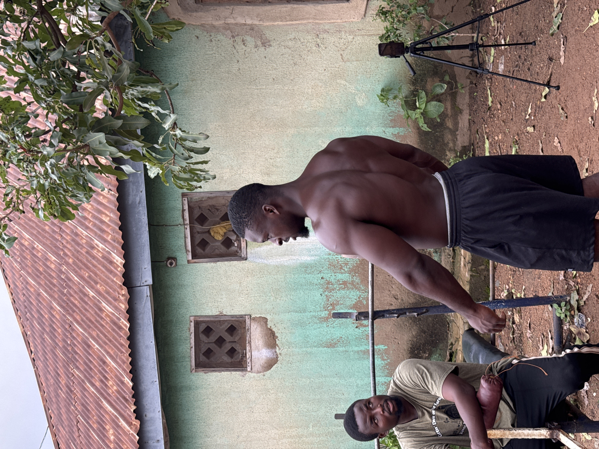 Two Black men are outdoors in a yard. One man, facing left, sits on a bench press, wearing a green t-shirt and black pants. He is looking to the left with his left hand resting on a weight. The other man, shirtless and wearing black shorts, faces away from the camera, looking to the left. Behind them is a light green building with a rusty corrugated metal roof. Two square windows with decorative grates are visible on the building. A tripod with a smartphone is set up to the right of the shirtless man, facing the building. The ground is dirt with some leaves scattered around. Lush green foliage is visible on the left and right sides of the scene. The overall scene suggests an informal outdoor workout area, possibly in a residential or communal space within Akwanga, Nigeria.