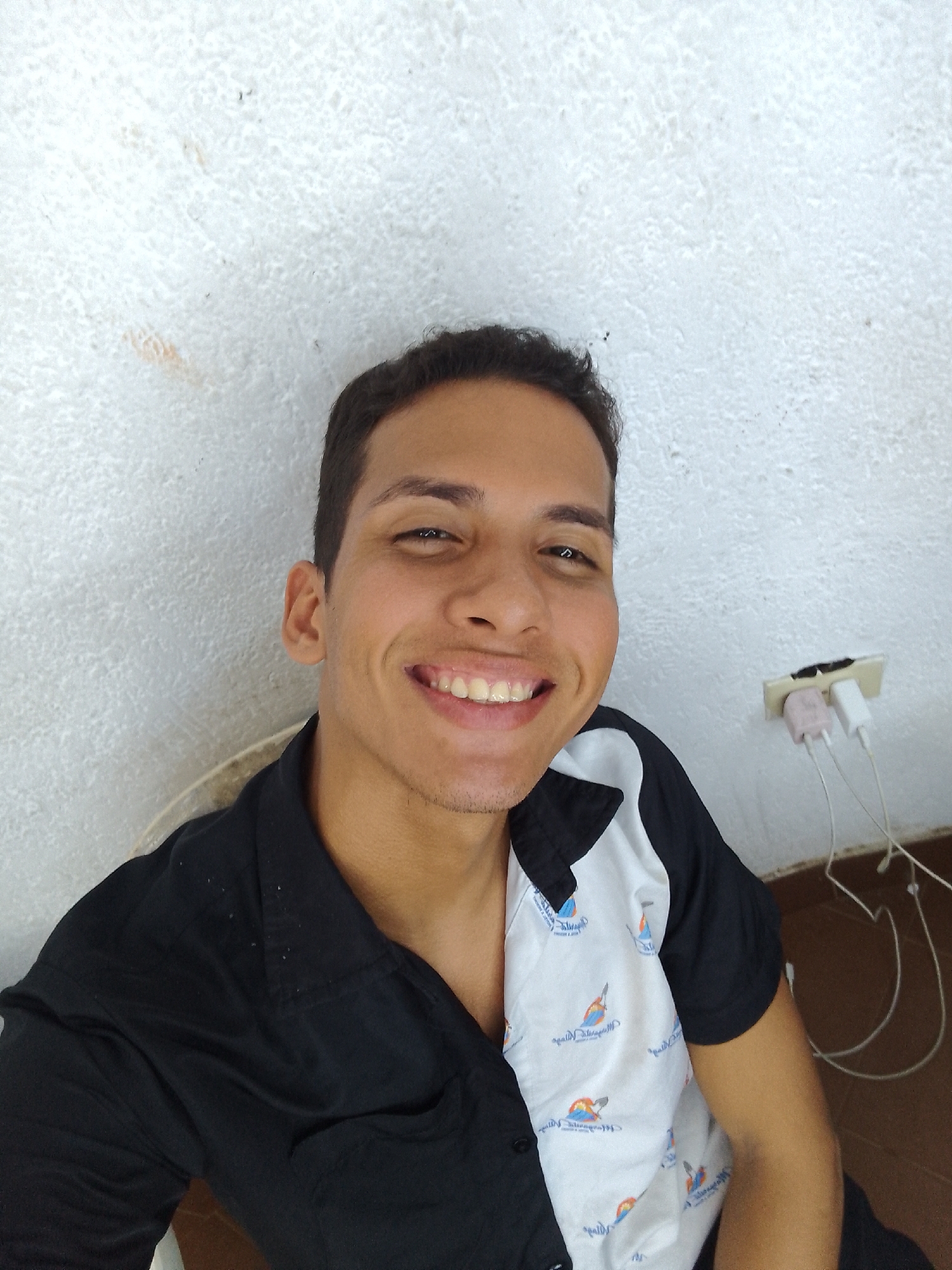 A light-skinned young adult male smiles at the camera in a selfie. He has short, dark hair, brown eyes, and a clean-shaven face. He is wearing a two-toned shirt, black on one side and white with bird illustrations on the other. Behind him is a white wall with a partially visible electrical outlet, which has two charging adapters plugged in, connected to wires. The floor is brown. Based on the man's appearance and the background, the image may be taken indoors, potentially in Porlamar, Venezuela.