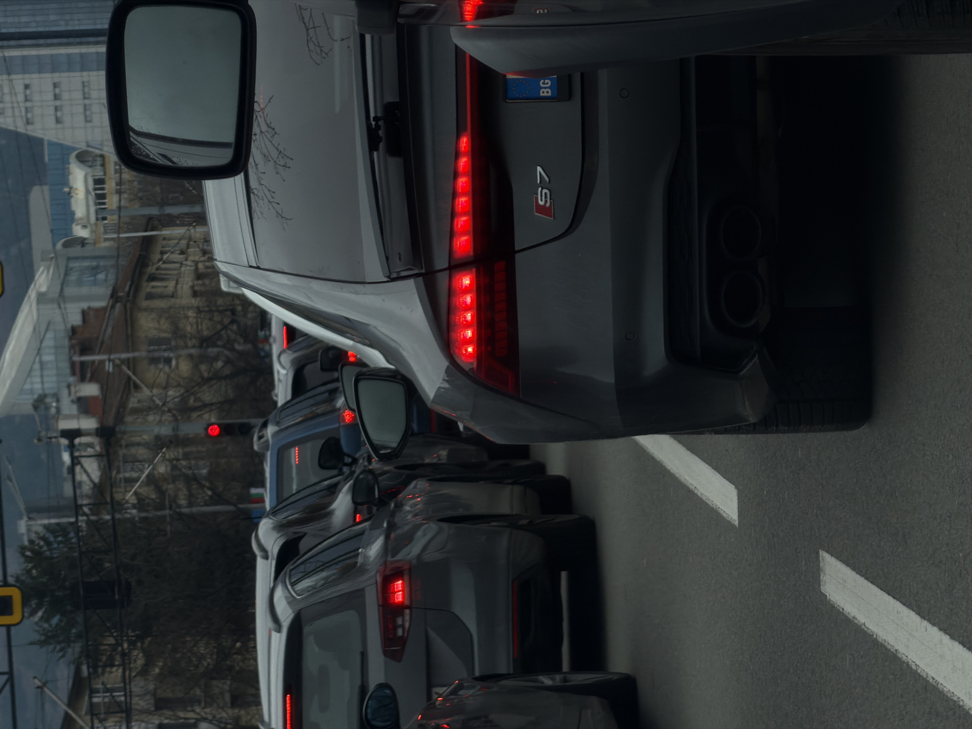 The image shows a line of cars stopped in traffic in Sofia, Bulgaria. The rear of a gray Audi S7 is prominent, with its illuminated taillights, "S7" badge, Bulgarian license plate, and exhaust pipes visible. The car is surrounded by other vehicles, with their taillights also illuminated. The street has white lane markings. Buildings and trees are visible in the background. A traffic light is visible. The image is shot at eye level.