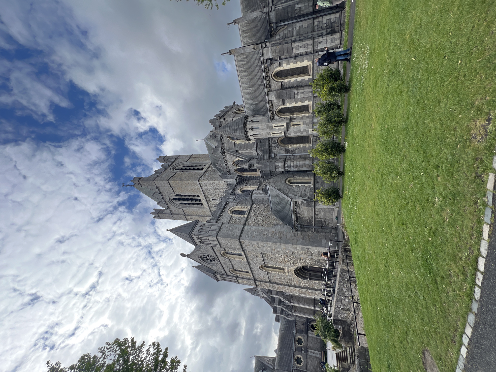 The image shows St. Patrick's Cathedral in Dublin, Ireland, as seen from a ground-level perspective. The cathedral, a large stone structure with multiple towers, occupies the central and right portions of the frame, contrasting with a blue and cloudy sky above. A patch of green grass extends along the right side of the image. A person is visible standing on a path next to the cathedral. Parts of the cathedral show architectural details such as arched windows, stone construction, and spires. The environment is urban, though partially obscured by the close-up view of the cathedral and greenery.