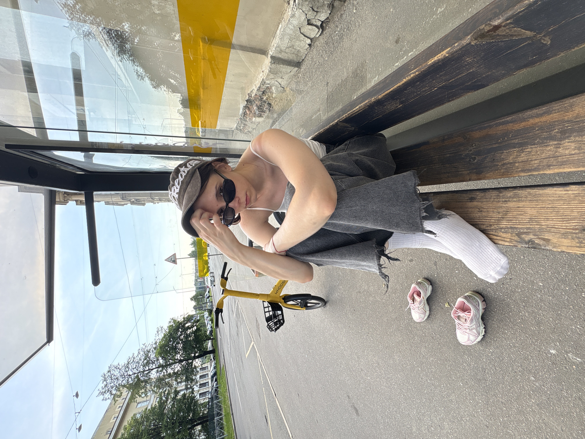 The image depicts a young woman sitting on a wooden bench at a bus stop in Saint Petersburg, Russia. She is wearing a white tank top, grey distressed jeans, white socks, sunglasses, and a baseball cap. Her left arm is resting on her knee, and her hand is touching her forehead, suggesting she might be contemplating or feeling unwell. Next to her on the pavement are a pair of pink athletic shoes. A yellow bike-sharing bicycle with a black basket is parked to her left. The bus stop has a glass enclosure and a yellow and white striped background visible through the glass. In the background, a street with cars, trees, and a building can be seen. The sky is partly cloudy, suggesting it is daytime. The overall scene conveys a sense of stillness and perhaps waiting.