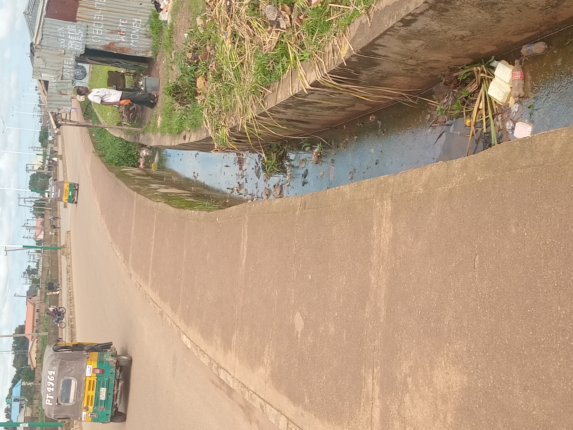 The image depicts a street scene in Bukuru, Nigeria. A three-wheeled vehicle, a "Keke Napep," with license plate PT 4964, is driving on the road. Beside the road, a concrete drainage channel is visible, filled with murky water and debris including plastic bottles and other refuse. A man in a white t-shirt and dark pants stands near a makeshift corrugated metal structure. The structure has writing on it, including "0803476," and "WHITE MONEY." He appears to be holding an orange object. Behind the man, there is a rusty chair. In the background, other buildings and trees are visible, along with streetlights and utility poles, suggesting an urban environment. The sky is overcast.
