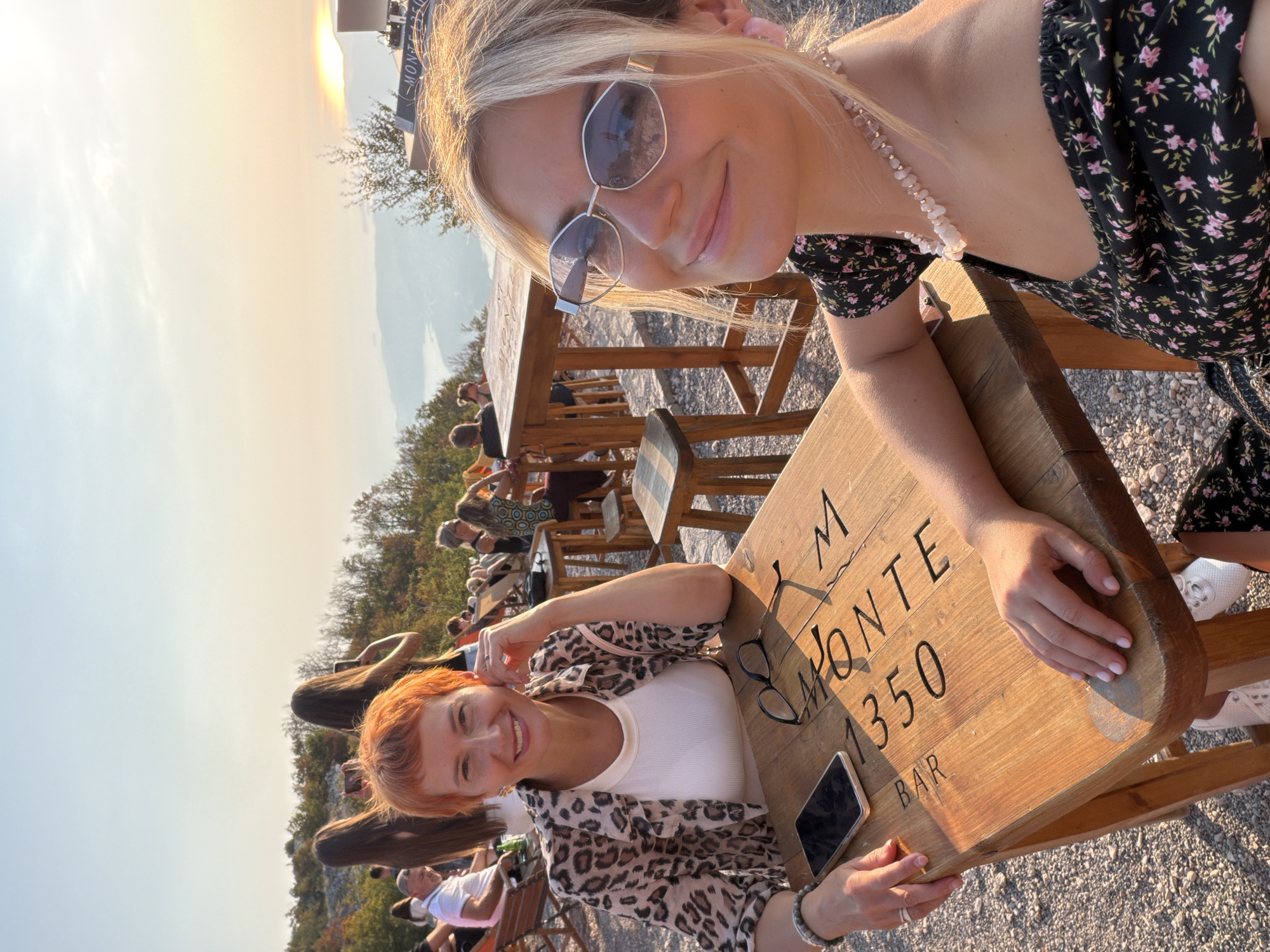 The image depicts an outdoor seating area at a bar located in Kuk, Montenegro, as evidenced by the text "MONTE 1350 BAR" visible on the wooden table. Two women are the primary subjects, seated at the table. The woman on the left, with short orange hair, wears a leopard-print jacket over a white top and is smiling at the camera, resting her hand on the table. Beside her is a smartphone and a pair of glasses. The woman on the right, with blonde hair and sunglasses, is also smiling and is closer to the camera, wearing a floral-print dress and a beaded necklace. The background shows a hazy mountainous landscape with a body of water, characteristic of the Boka Bay region, under a warm, sunset sky. Other patrons are visible in the background, seated at tables and on chairs. The seating area is paved with gravel.