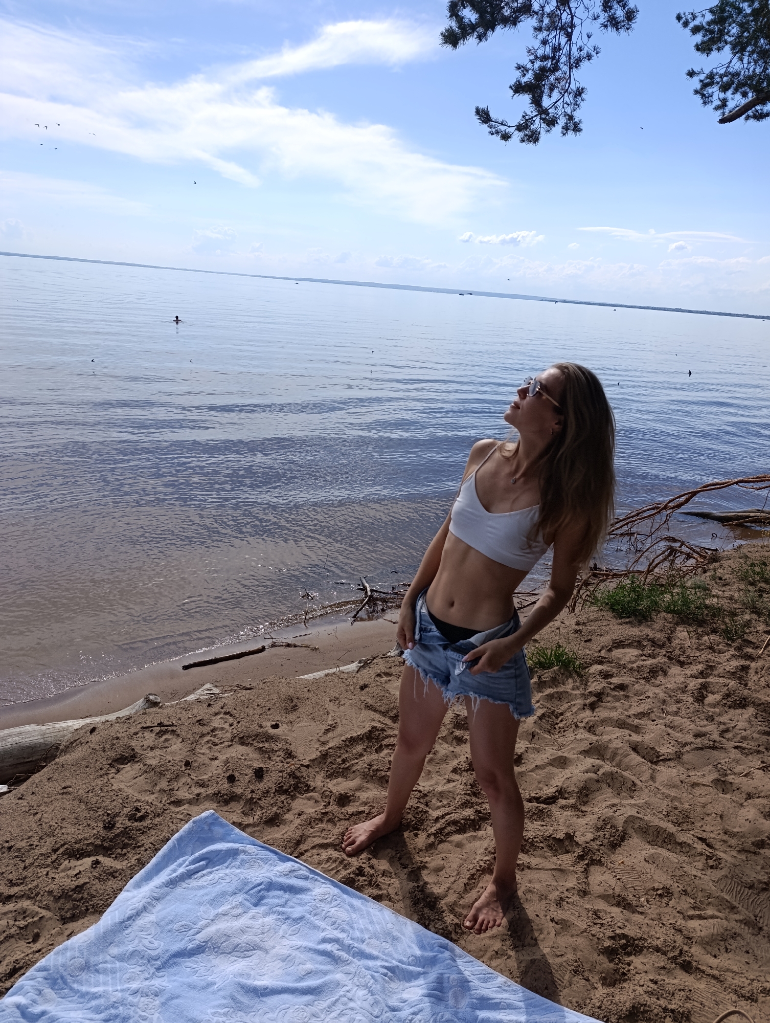 This is a full-body shot of a young woman standing on a sandy beach next to a body of water. She is wearing a white bikini top and denim shorts, with her hands on the waistband of the shorts. The woman is looking up and to her right, with a slight smile on her face, suggesting a relaxed and happy mood. Her long, blonde hair is down and slightly windswept.
The background consists of a large expanse of water, with a faint shoreline visible in the distance. The sky is blue with scattered white clouds, indicating it is daytime, likely late morning or early afternoon, under fair weather conditions. There are a few birds flying in the sky and a small boat on the water. Some tree branches hang into the frame from the top right, suggesting the beach is near a wooded area. A light blue towel is spread out on the sand in the foreground. The scene evokes a sense of leisure and enjoyment of a summer day by the water.