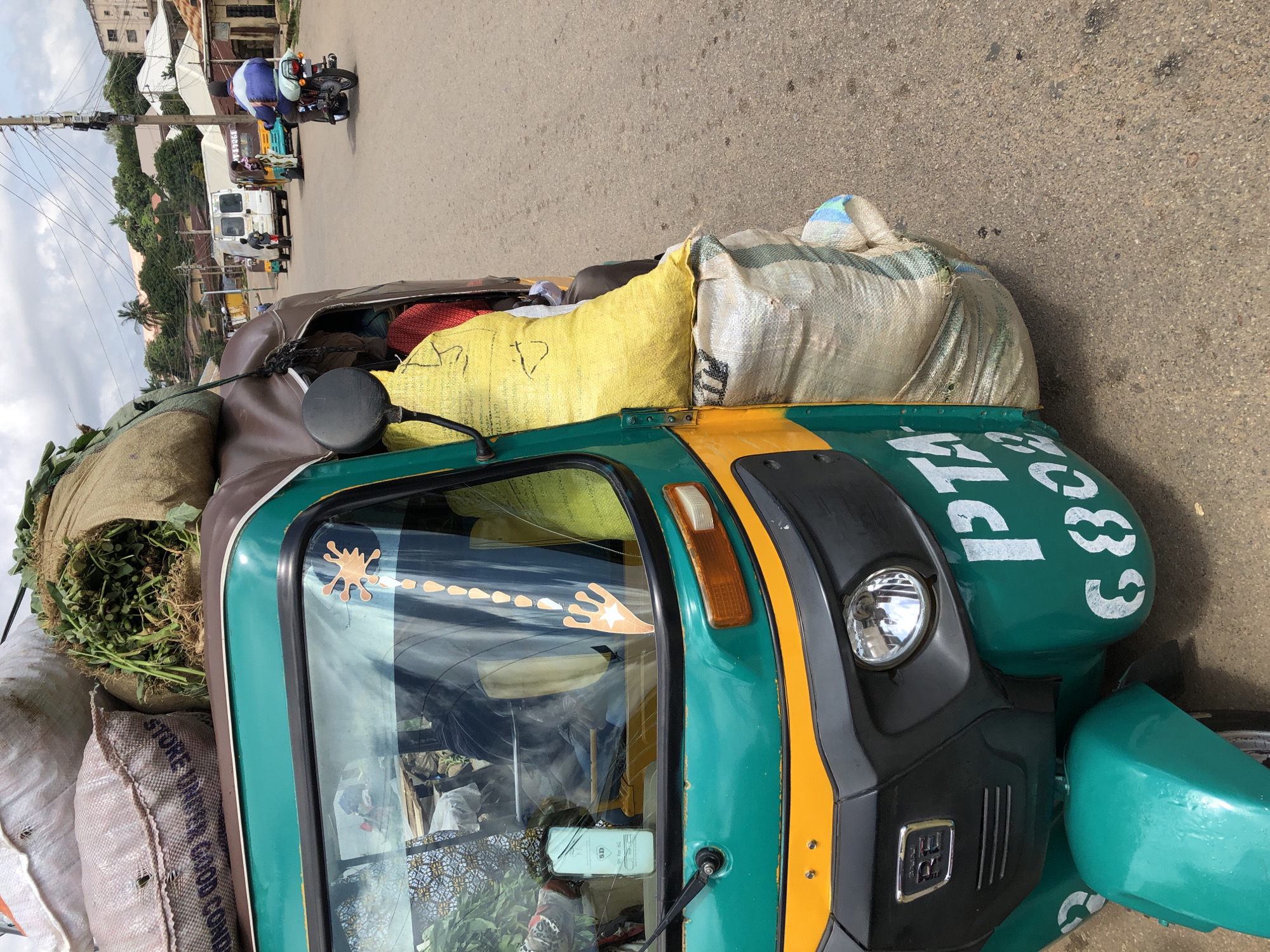 The image shows a three-wheeled auto-rickshaw heavily laden with sacks and bundles of what appears to be produce and possibly other goods. The auto-rickshaw is predominantly green with yellow accents and has white lettering that reads "PTA" and "6802" on its side. The vehicle is on a paved road in an urban setting. In the background, there are buildings, trees, and utility poles with numerous wires. Other vehicles, including a white van and a motorcycle with a rider, are visible on the road. The sky is partly cloudy. The context suggests a scene in Jos, Nigeria, likely a marketplace or a street where goods are transported.