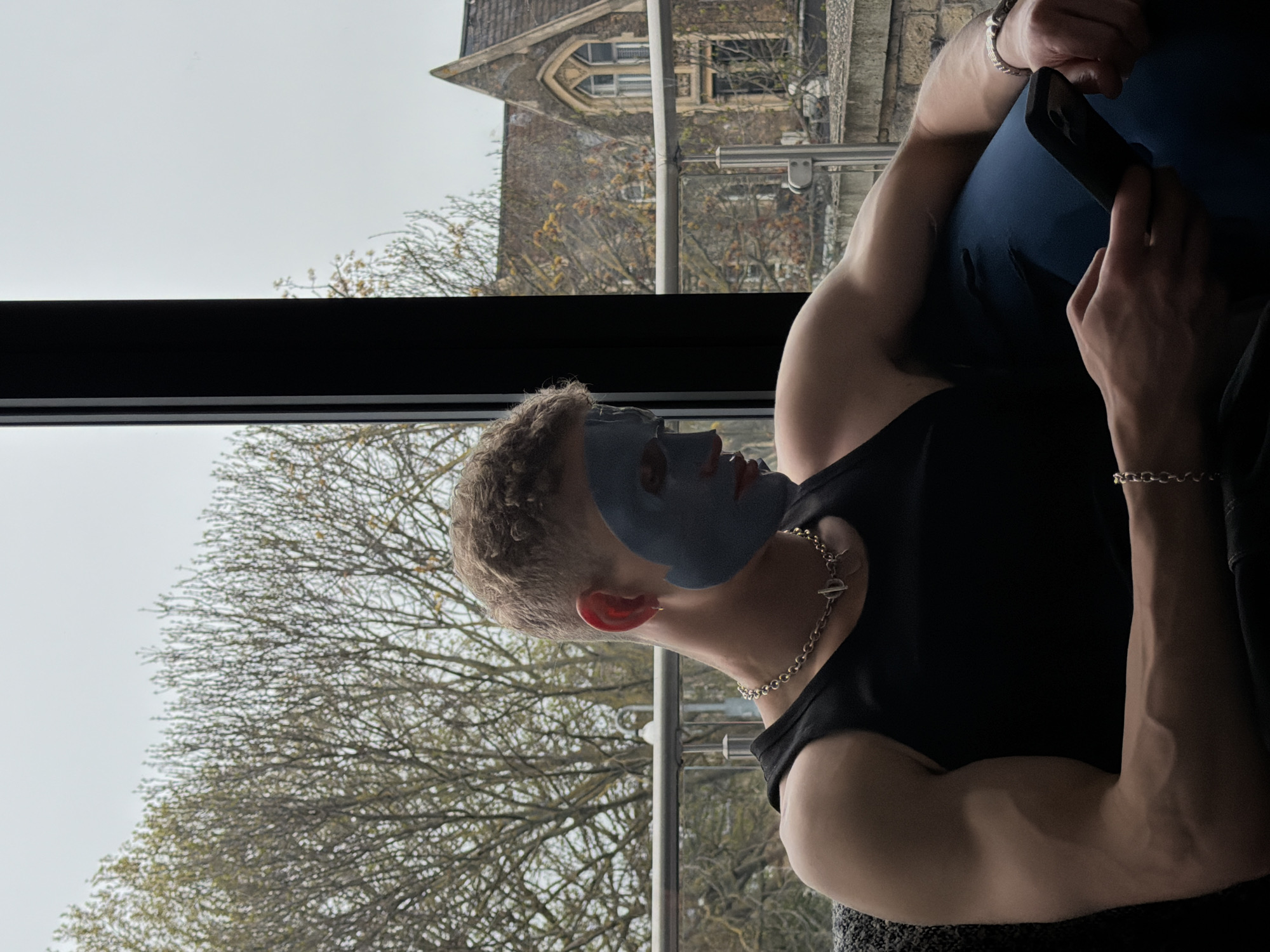 A man sits, facing right, in front of a window, wearing a black tank top, jewelry, and a blue facial mask. He holds a cell phone in his left hand. Behind him, visible through the window, is a tree with bare branches and a stone building. This is a view of the back of St. Paul's Cathedral Choir School in London, United Kingdom.