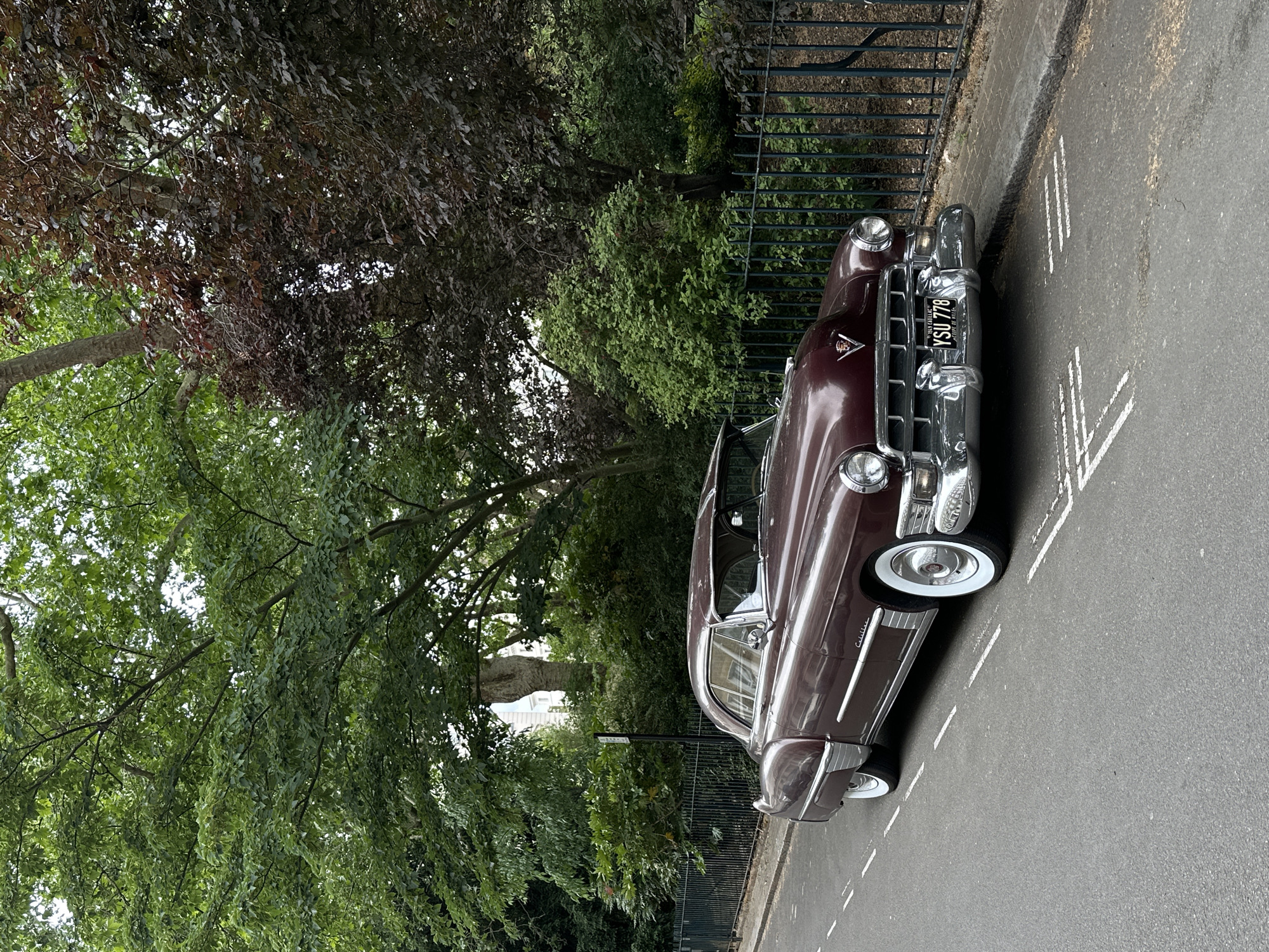 A vintage dark-red Cadillac automobile is parked on a street in London, United Kingdom. The vehicle is positioned on the right side of the road with white painted lines. A metal fence runs parallel to the car, separating the street from a green area filled with trees and shrubs. The car's front end and license plate are prominently visible. The license plate reads "YSU 778". The setting suggests a residential or park area within London.