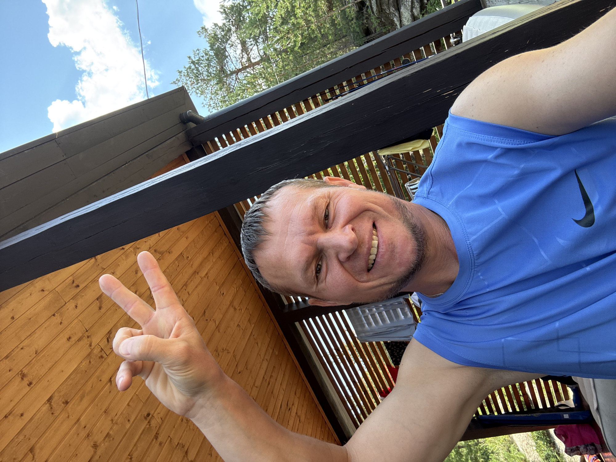 A mid-shot selfie shows a man with short brown hair and stubble, smiling at the camera. He is wearing a blue sleeveless t-shirt with a Nike logo. His right arm is raised, and his hand is held up with his index and middle fingers extended in a V-shape, while his thumb, ring finger, and pinky finger are tucked in. He is standing outdoors, in front of a wooden structure with a black beam and a slatted wall. In the background, trees and a rocky outcrop are visible under a blue sky with white clouds. The setting appears to be a recreational area or a residential property, possibly a dacha or cabin, given the rustic wooden construction. The specific location of New Bukhtarma, Kazakhstan is not evident from the image itself, but the environment suggests a natural, possibly mountainous or forested, region.