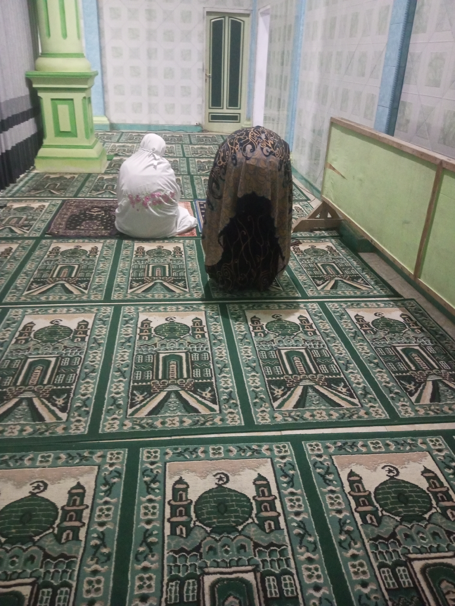 In the city of Andongsili, Indonesia, two individuals are observed engaged in prayer within what appears to be a prayer room or mosque. The floor is covered with prayer mats featuring a repeating pattern of green mosque designs. To the left, a person wearing a white hijab is kneeling, facing away from the camera. To their right, another person is also kneeling, shrouded in a patterned dark brown and tan cloth that covers their head and body. The room's walls are decorated with a repeating light blue and white geometric pattern. A prominent green column stands to the left, and a green wooden partition is visible on the right. A closed door with green trim is situated in the background.