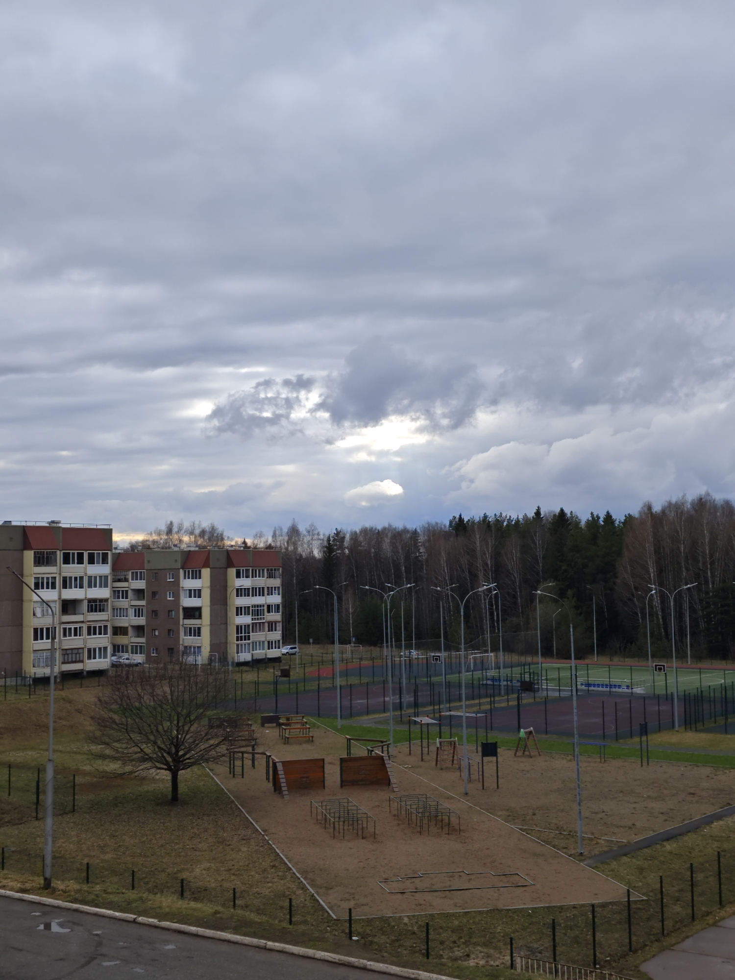 This elevated wide shot captures an urban and natural landscape under a heavily overcast sky in Markovskii, Russia. The scene unfolds during daytime, characterized by muted light piercing through dense clouds, creating bright patches on the horizon. The overall impression is one of quiet, with no people visible.
In the foreground and midground, a diverse recreational area is prominent. A bare, deciduous tree stands on a patch of sparse, brownish grass. Adjacent to this, a sandy outdoor workout area features various metal exercise structures like parallel bars and monkey bars, along with some wooden obstacle course elements. Fences, including chain-link and simple metal posts, delineate these spaces. A section of a dark, paved surface, possibly a road or parking lot, with small puddles, is visible in the bottom left corner.
Beyond the workout zone, a series of sports courts are laid out. These include a football pitch with goalposts and a basketball court with hoops, both enclosed by tall fences and featuring artificial turf or similar surfacing in green and reddish-brown hues. Numerous tall lampposts are strategically placed around these facilities and along the edge of the forest.
On the left side of the frame, two multi-story apartment buildings dominate, featuring beige or light yellow facades with red roofs. Their architecture appears typical of residential blocks found in the region. In the background, a dense forest stretches across the horizon, comprising both leafless deciduous trees and evergreen conifers, indicating a cooler season such as late autumn or early spring. No visible text is present in the image.