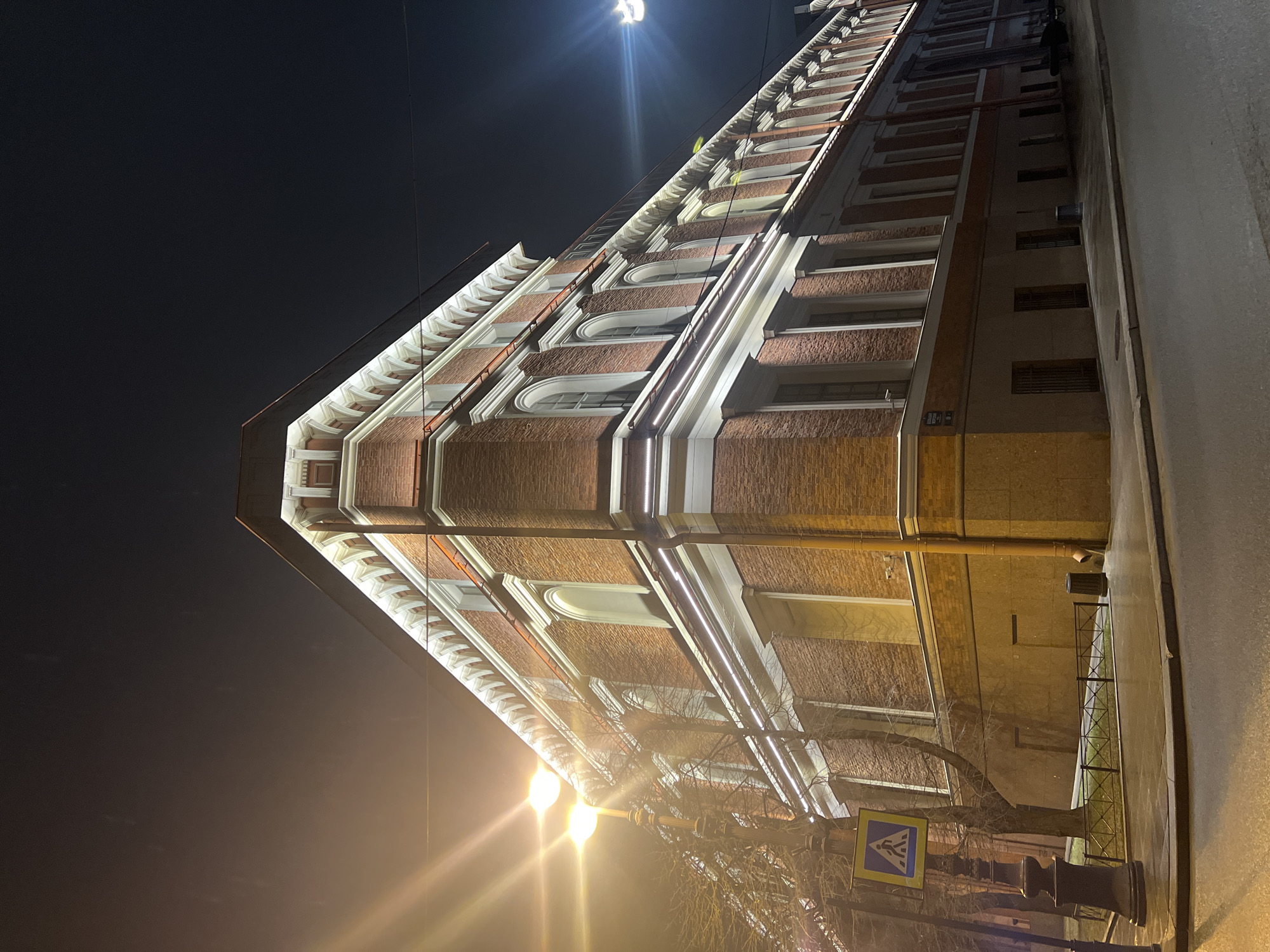 The image displays the exterior of an illuminated corner building located at Gorokhovaya Street, 8, Saint Petersburg, Russia, captured at night from a low-angle perspective. The multi-story structure features a red brick facade with white architectural trim, including prominent cornices, string courses, and window surrounds. The two lowest visible floors are clad in a dark grey stone, with rectangular windows fitted with metal grates.
Linear LED lighting strips are integrated along the cornices and below the window lines, providing uniform cool white illumination to the architectural details. The upper floors present a series of regularly spaced windows, many exhibiting arched tops.
In the foreground to the left, a dark, ornate lamppost emits a bright, warm orange light, creating a lens flare in the upper left quadrant. A bare-branched deciduous tree stands behind the lamppost. A blue and yellow pedestrian crossing sign, depicting a walking figure, is affixed to the lamppost's pole.
The street in the foreground consists of wet asphalt, indicating recent precipitation or dampness, and curves gently to the right. A paved sidewalk borders the building, separated from the lower facade by a low, dark metal fence enclosing a narrow strip of grass. A dark, cylindrical trash bin is situated on the sidewalk near the fence. A second bright light source, likely another streetlight, is visible in the upper right background. The night sky is dark and featureless. No individuals, vehicles, or observable actions are present within the scene.