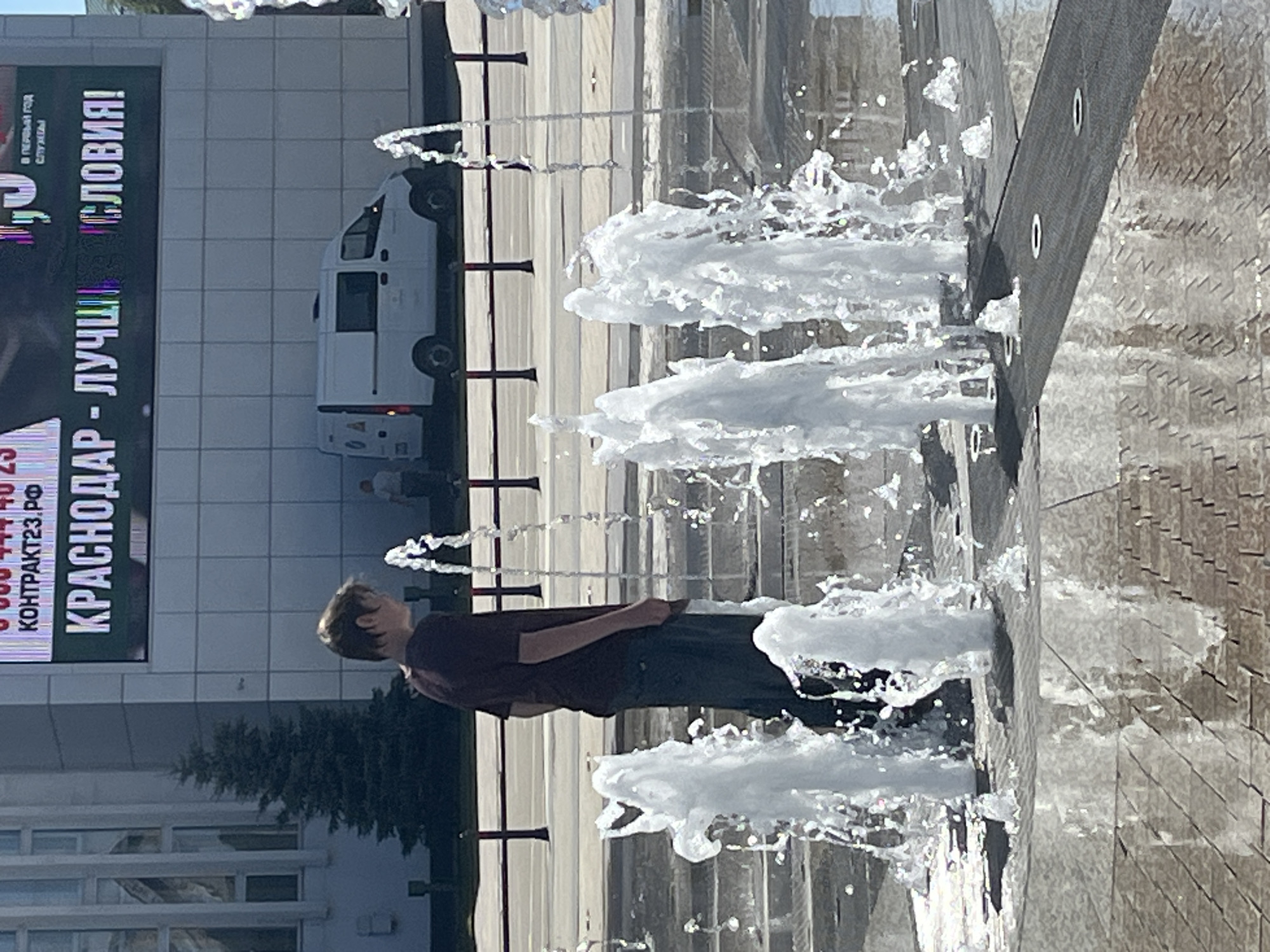 In Krasnodar, Russia, a teenage boy stands partially obscured by a fountain, facing away from the camera. He wears a maroon t-shirt and dark pants. The fountain's water jets create a dynamic, abstract foreground. In the background, a white van is parked in front of a modern building adorned with a large digital billboard displaying text, including "KRASNODAR - LUCHSHIY" in Russian. The scene captures a bright, urban environment with a playful interaction with a water feature.