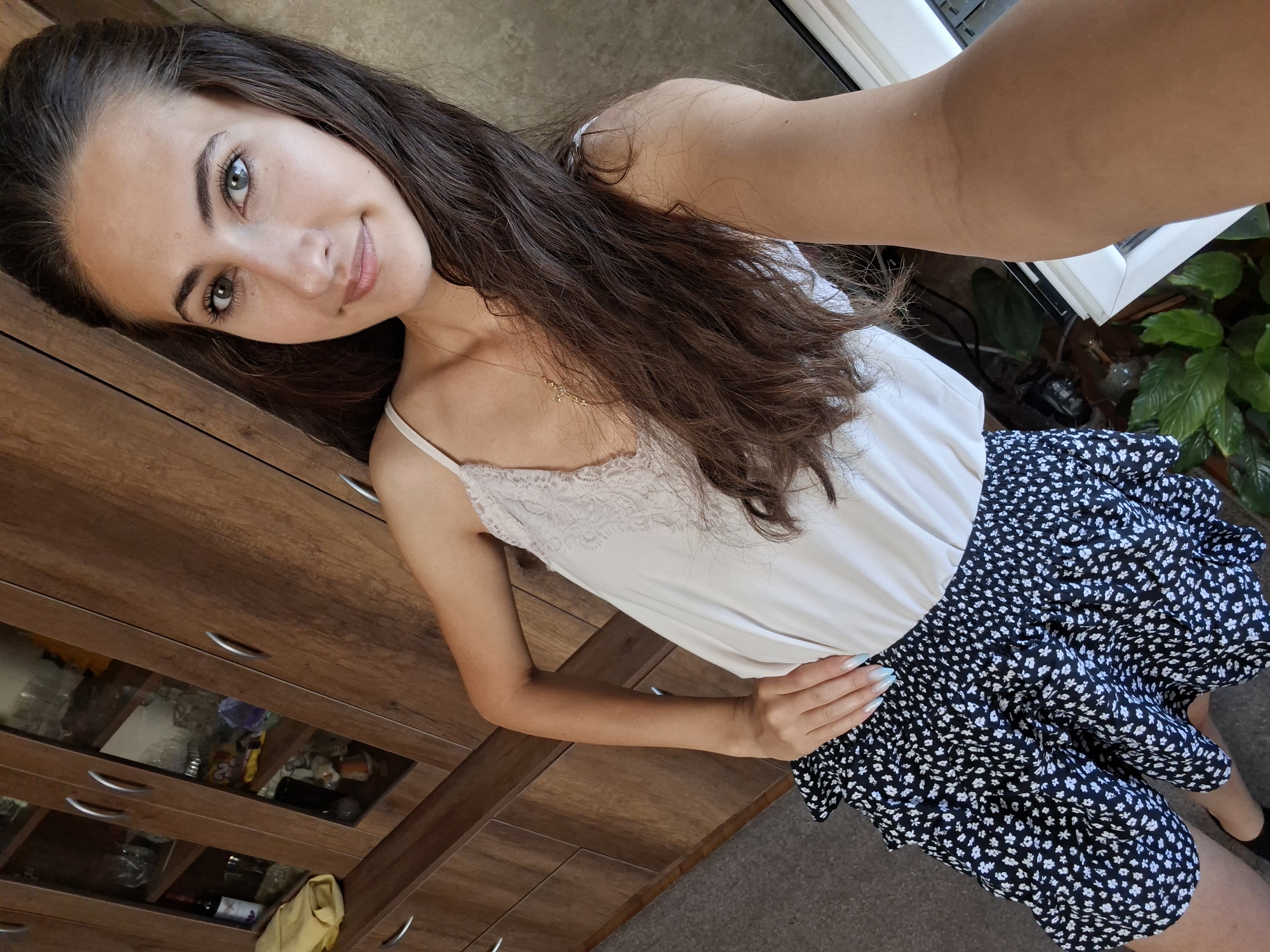 In an indoor setting, a young woman with long, dark brown hair and green eyes is taking a selfie. She is wearing a white camisole with lace trim and a black and white floral print skirt. Her right hand is on her hip, and her left arm is resting on her thigh. She is standing in front of a wooden cabinet with glass doors. The cabinet contains various items, including glassware and decorative objects. A yellow cloth is visible on one of the shelves. The floor is carpeted. There are also some potted plants visible near a window. The lighting appears to be natural, coming from the window.