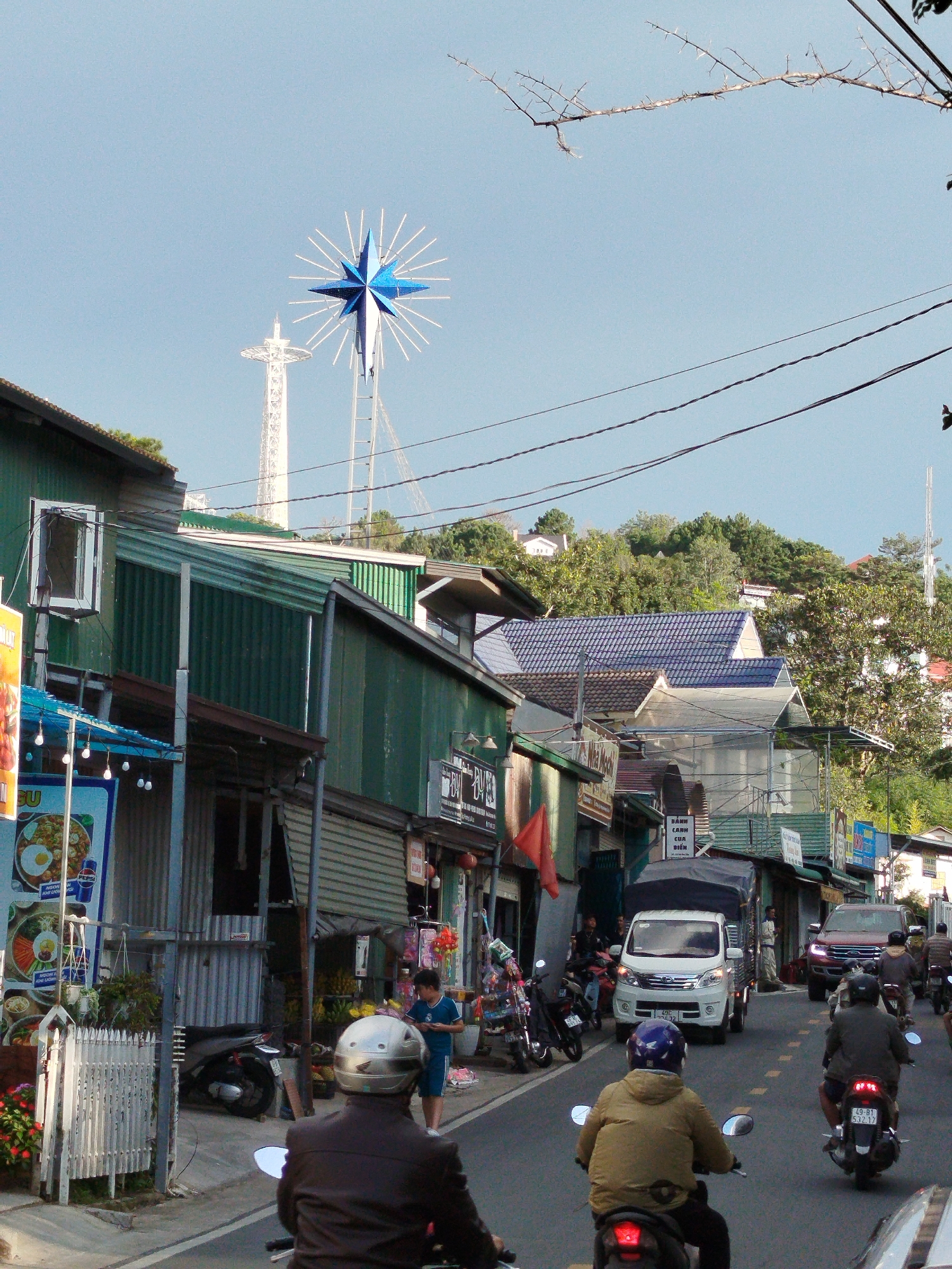 In Da Lat, Vietnam, a street scene unfolds under a clear sky, dominated by a striking blue and white star-shaped structure atop a tower in the distance. The foreground features a man on a motorcycle, wearing a silver helmet and a brown jacket, facing away from the camera. To his right, another motorcyclist, identified by a blue helmet and a tan jacket, is also moving away. Several other motorcycles are parked on the left side of the street, and a white truck is visible further down.
The street is lined with buildings constructed from corrugated metal, painted in shades of green and gray. Some of these buildings have open doorways and windows, revealing glimpses of what appear to be shops or residences. A white fence runs along the left side of the street, bordering a small patch of flowering plants. In the background, a white tower with a distinctive shape, possibly an observation tower, stands tall, while lush green trees and distant buildings cover the hillside. The overall impression is of a busy, developing urban environment, characteristic of a Vietnamese city like Da Lat.